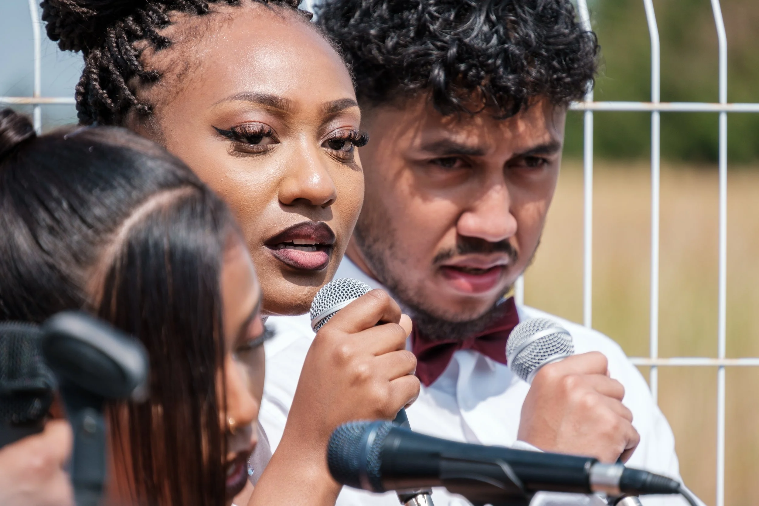 Trois jeunes personnes tenant des microphones lors d'une réunion ou d'un discours en plein air, avec un fond de grillage métallique.