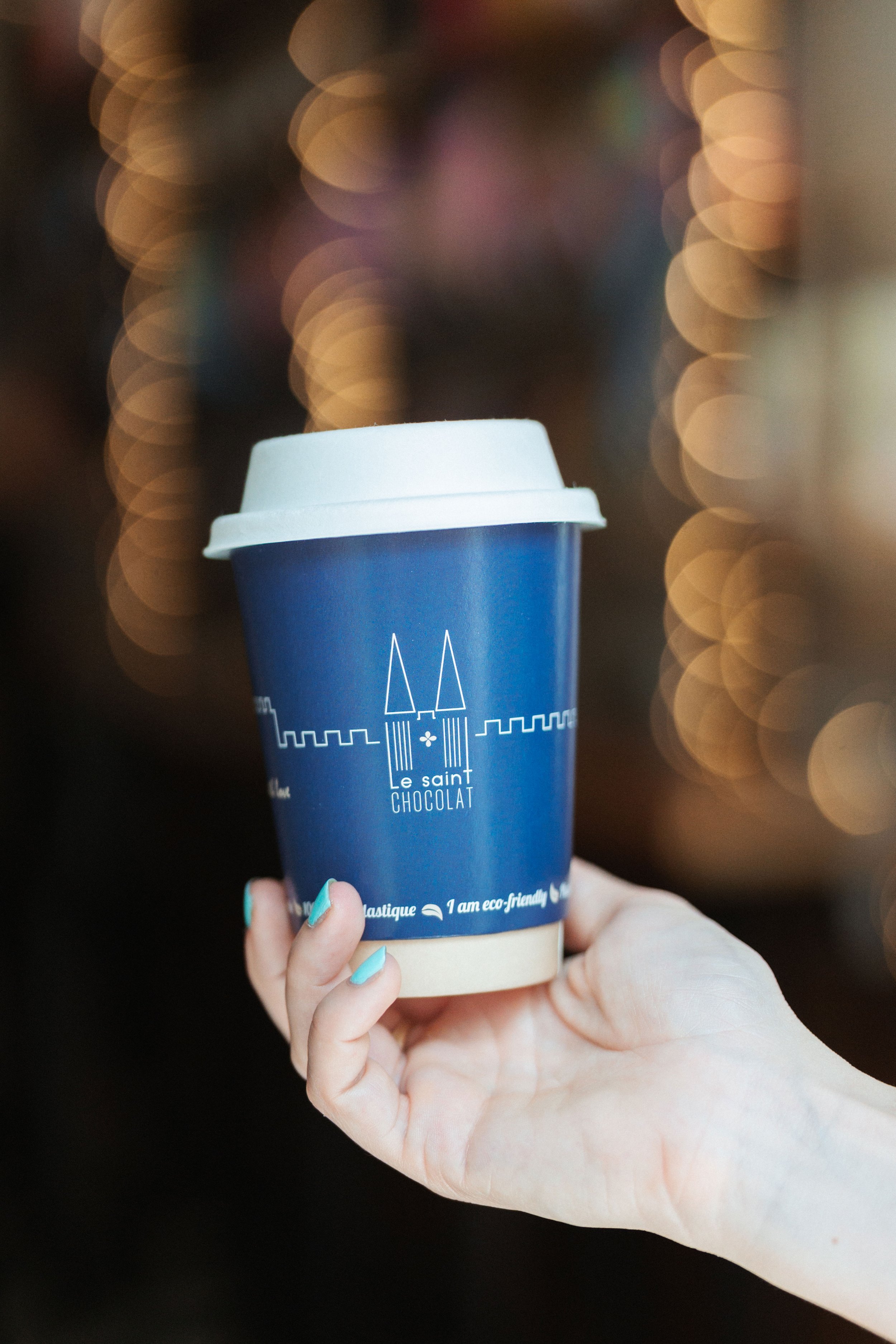 Une main tenant un gobelet en carton bleu avec un couvercle blanc, portant le logo "Le saint CHOCOLAT" et des illustrations de tours, contre un fond flou avec des lumières en cercle.