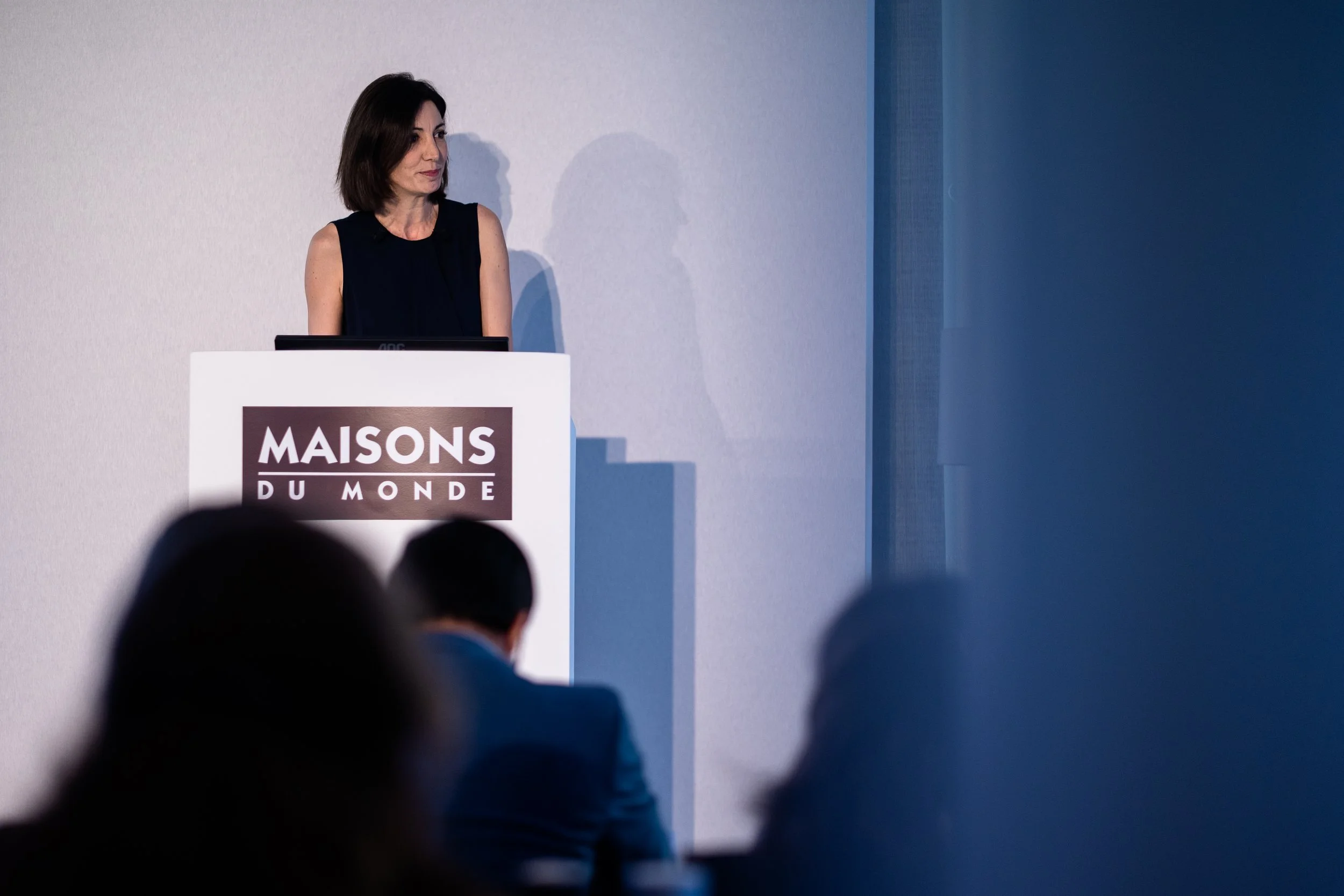 Une femme se tient derrière un pupitre avec le logo 'Maisons du Monde', lors d'une présentation ou conférence, avec quelques personnes dans le public en silhouette en premier plan.