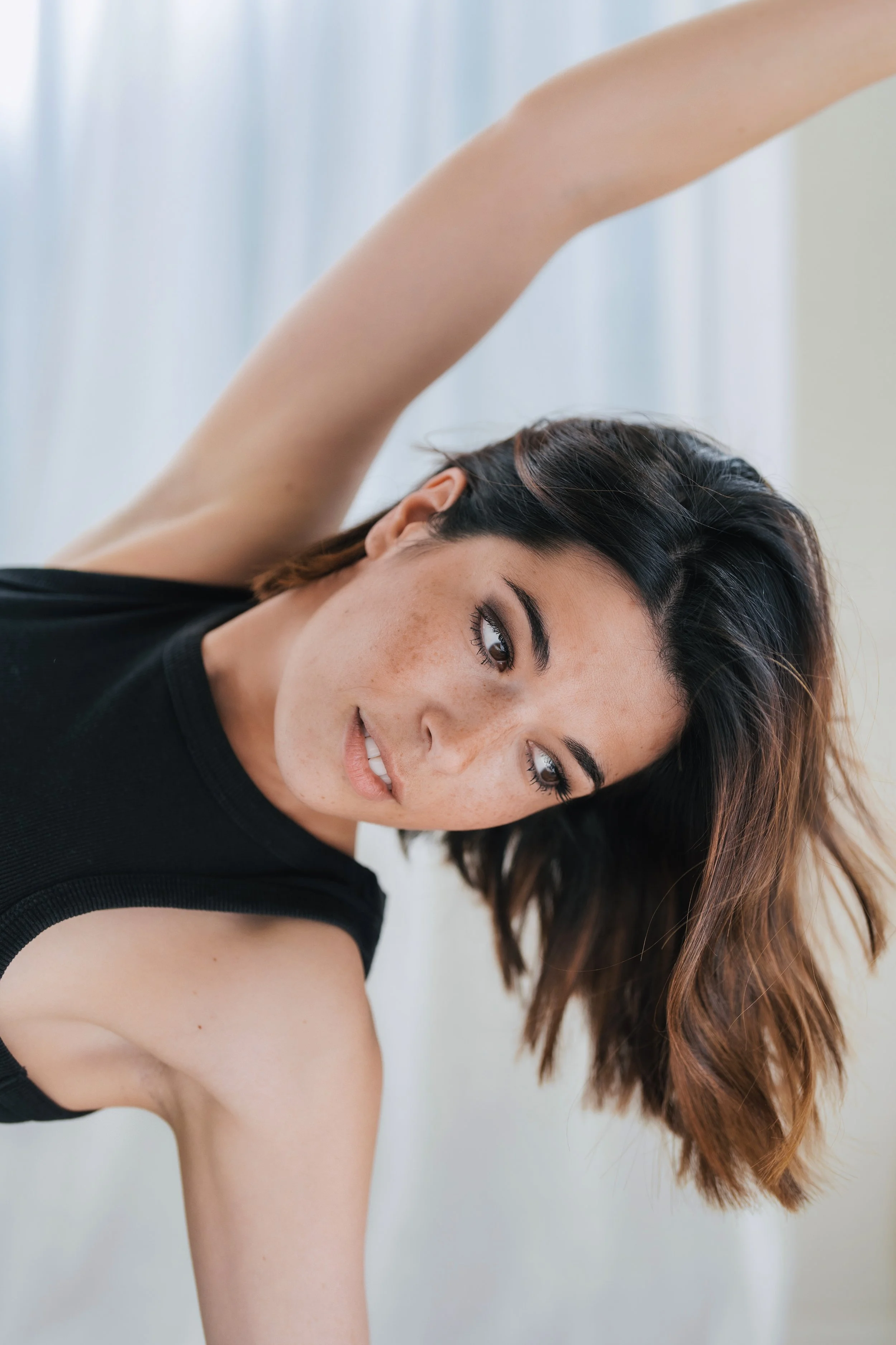 Femme faisant du yoga, étirée avec un bras au-dessus de la tête, portant un débardeur noir, visage souriant, cheveux bruns ondulés, fond léger et lumineux.