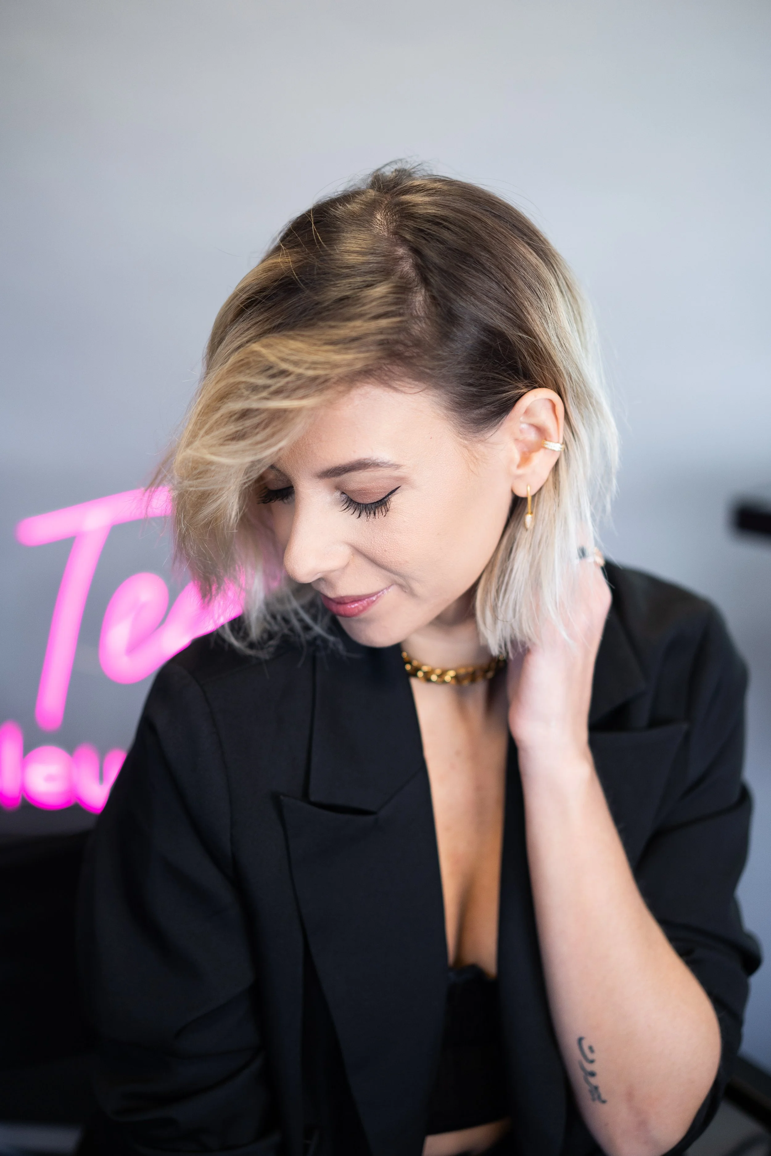 Une femme avec des cheveux blonds, coiffées de manière asymétrique, porte un blazer noir et des bijoux en or, regardant vers le bas, avec un fond flou et un panneau de néon rose.
