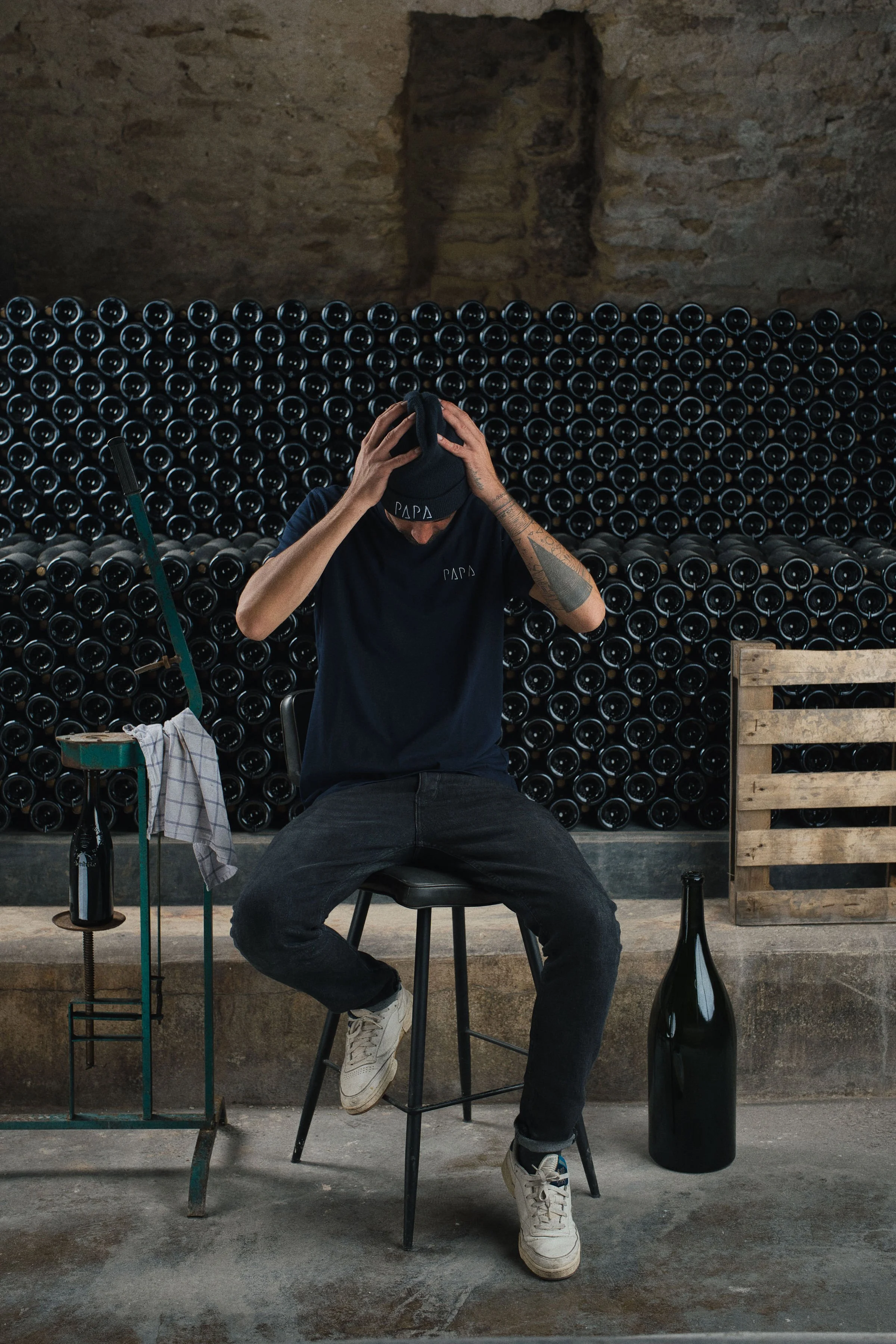 Homme assis sur un tabouret, portant un t-shirt noir avec 'PAPA' écrit dessus, tenant une casquette sur la tête, avec une cave à vin en arrière-plan.