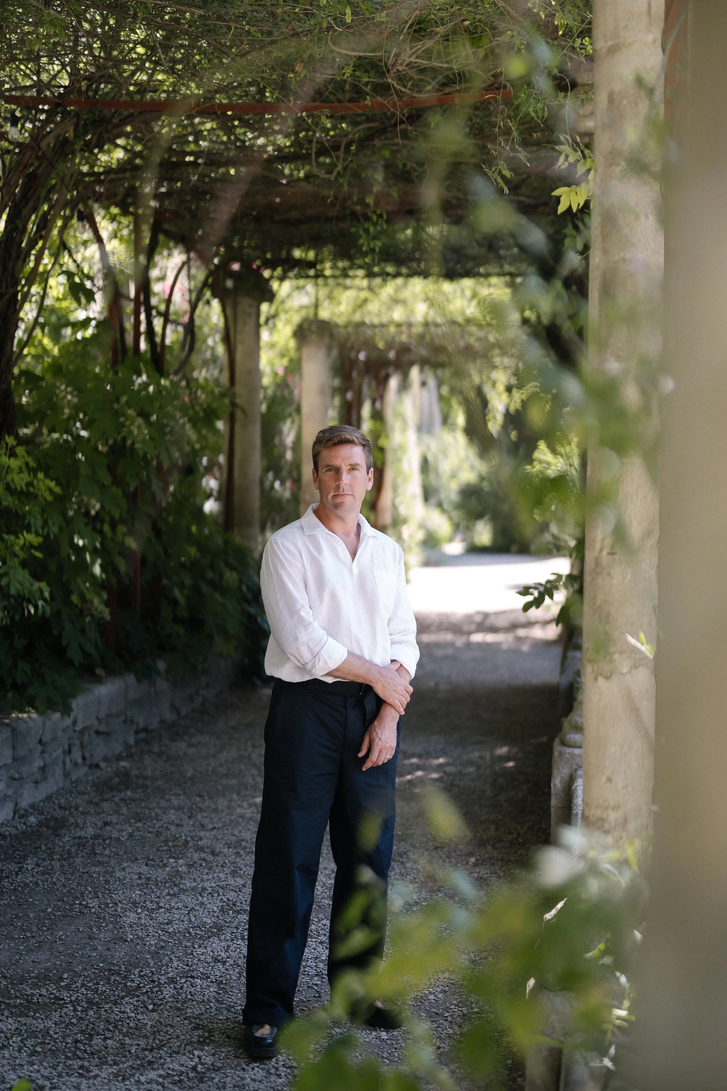 Un homme en chemise blanche et pantalons noirs se trouve dans un sentier entouré de verdure et une structure en pierre, avec des colonnes et une pergola recouverte de plantes.
