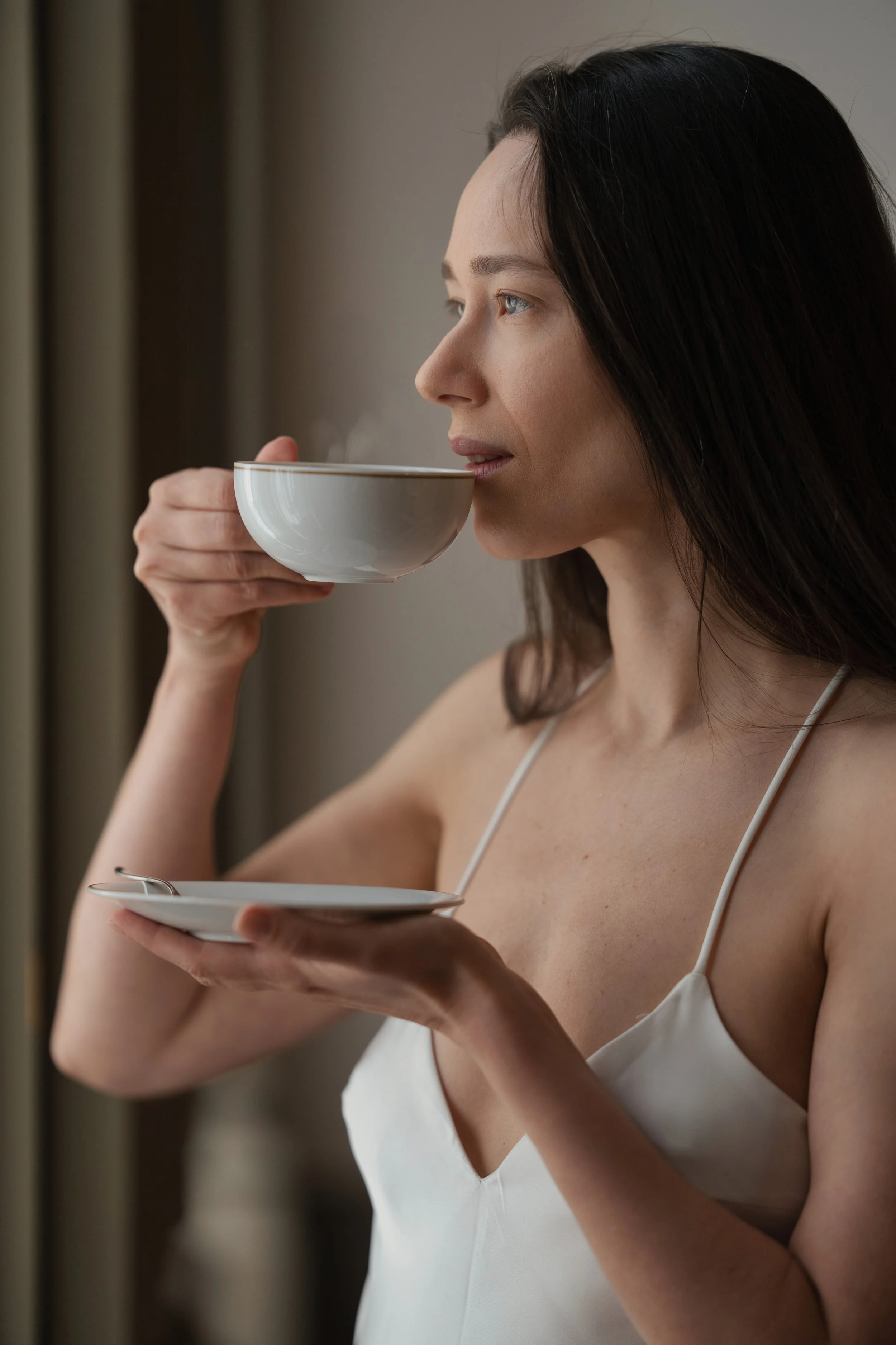 Femme prenant une tasse de café ou de thé, vue de profil, portant une robe blanche, dans un intérieur calme.
