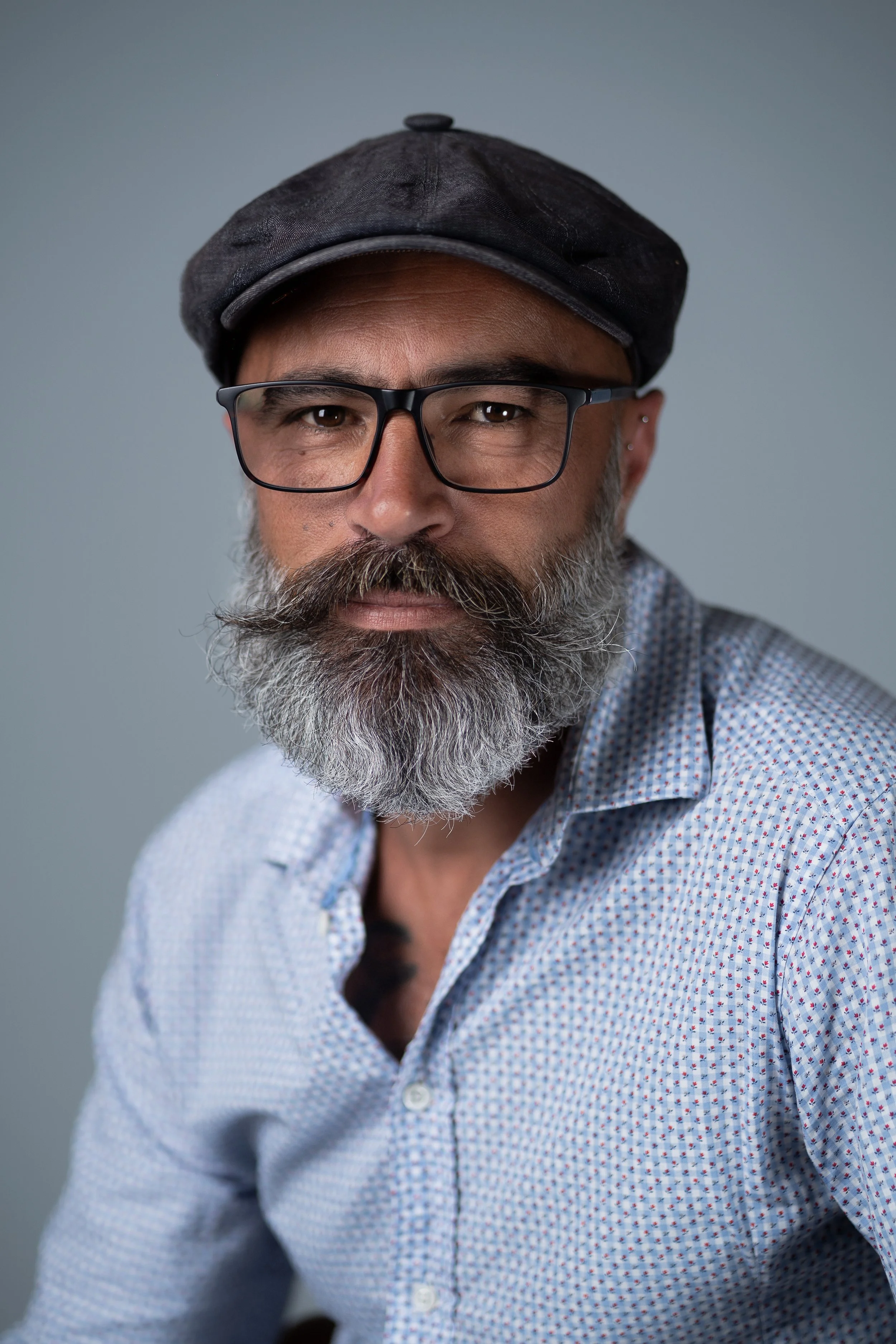 Portrait d'un homme barbu portant des lunettes et un béret noir, habillé d'une chemise à petits motifs, contre un fond gris.