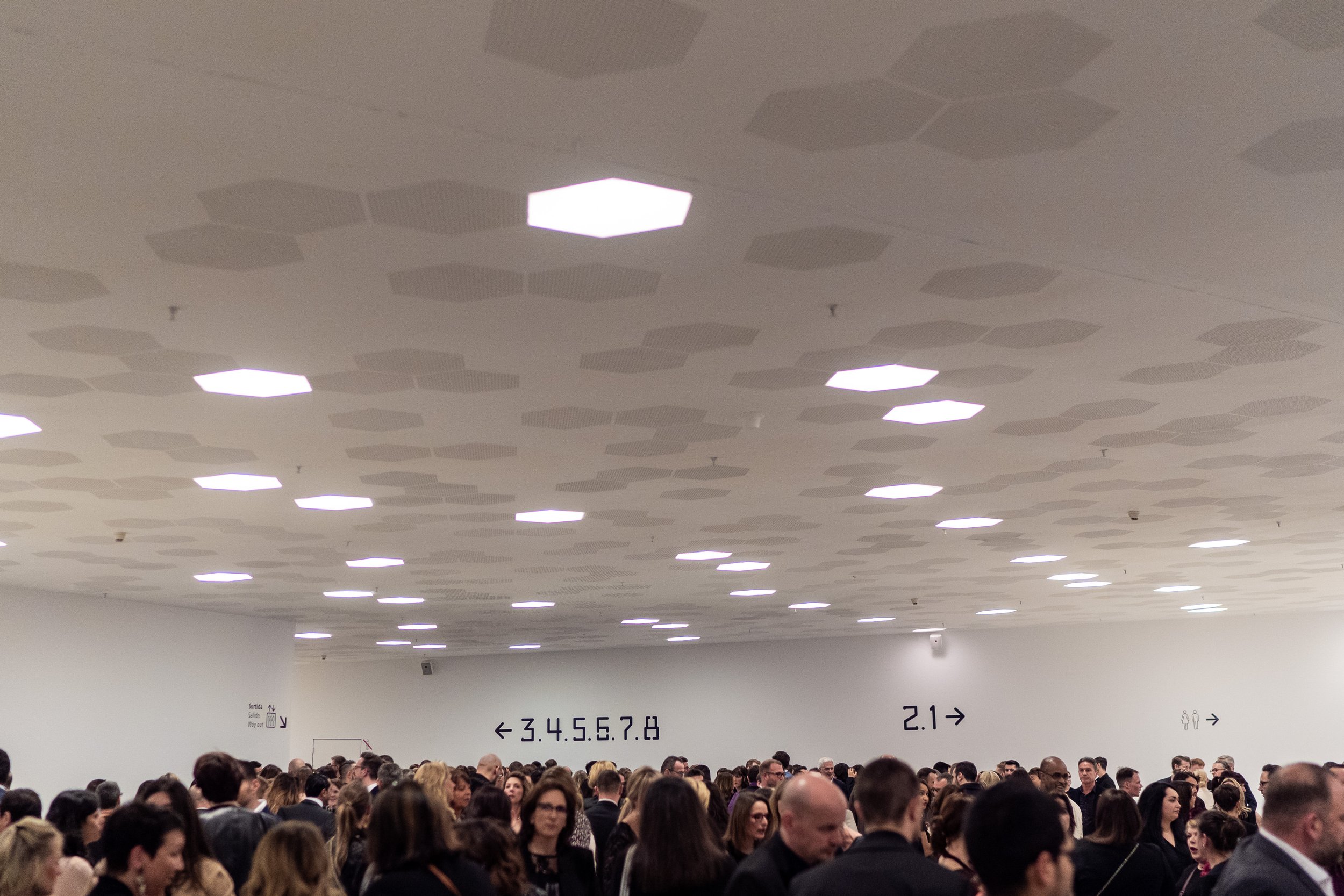 Salle d'attente ou de transit bondée avec de nombreuses personnes, murs blancs avec des indications en japonais et en chiffres, plafond avec motifs géométriques.