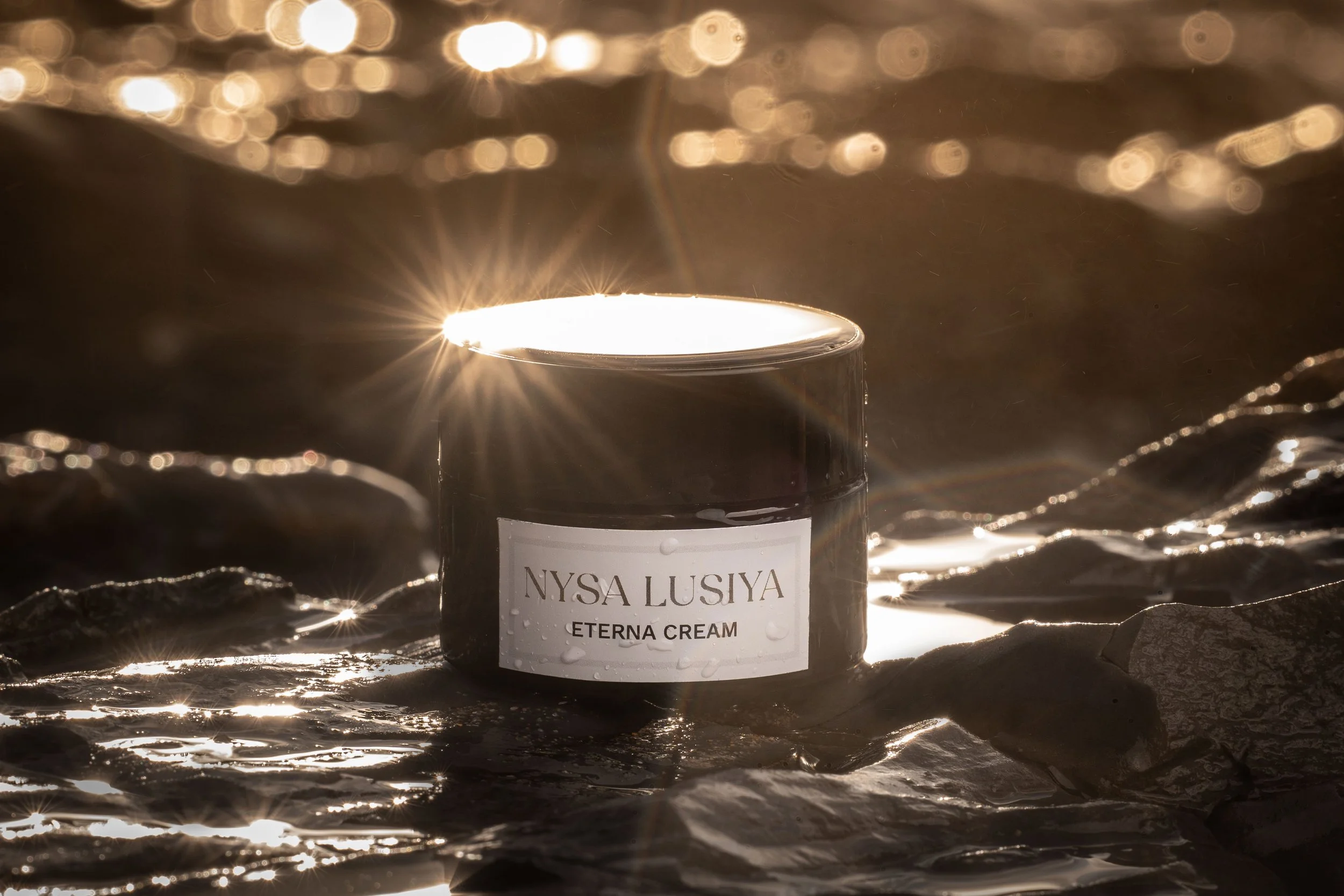 Un pot de crème nommé NYSA LUSIYA ETERNA CREMA, placé sur des rochers au bord de l'eau, avec la lumière du soleil réfléchie sur la surface du pot et sur l'eau environnante.