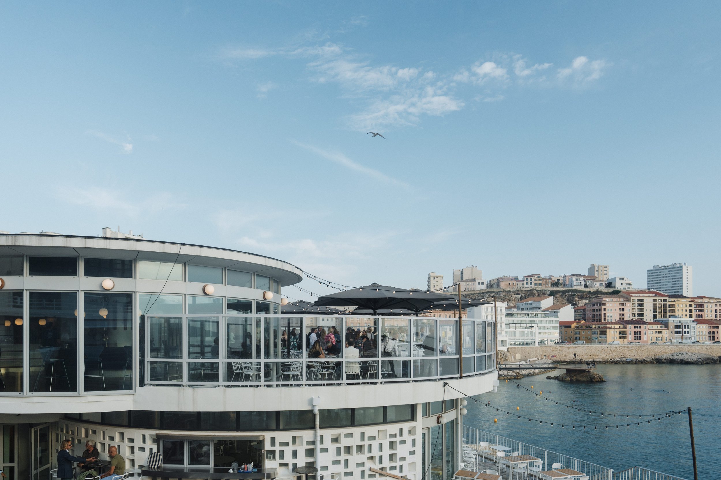 Un restaurant en bord de mer avec des personnes à l'intérieur et sur la terrasse, un ciel bleu avec quelques nuages, et une ville en arrière-plan.