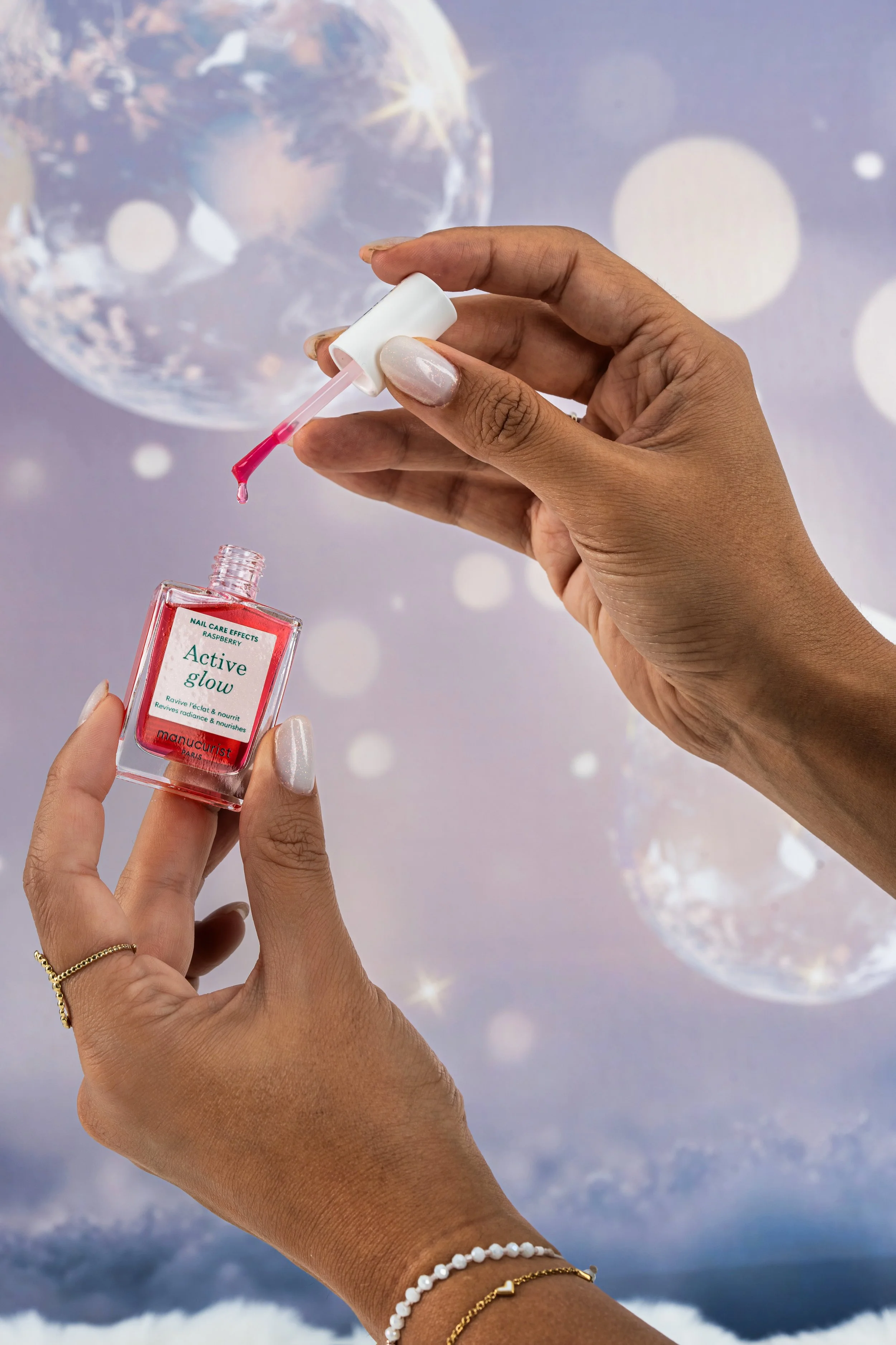 Main tenant un vernis à ongles rose avec une pince à ongles, tenant un flacon de vernis à ongles rouge nommé "Active glow" de la marque Manucurist, contre un fond flou avec des décorations en forme de boules brillantes et de lumières.