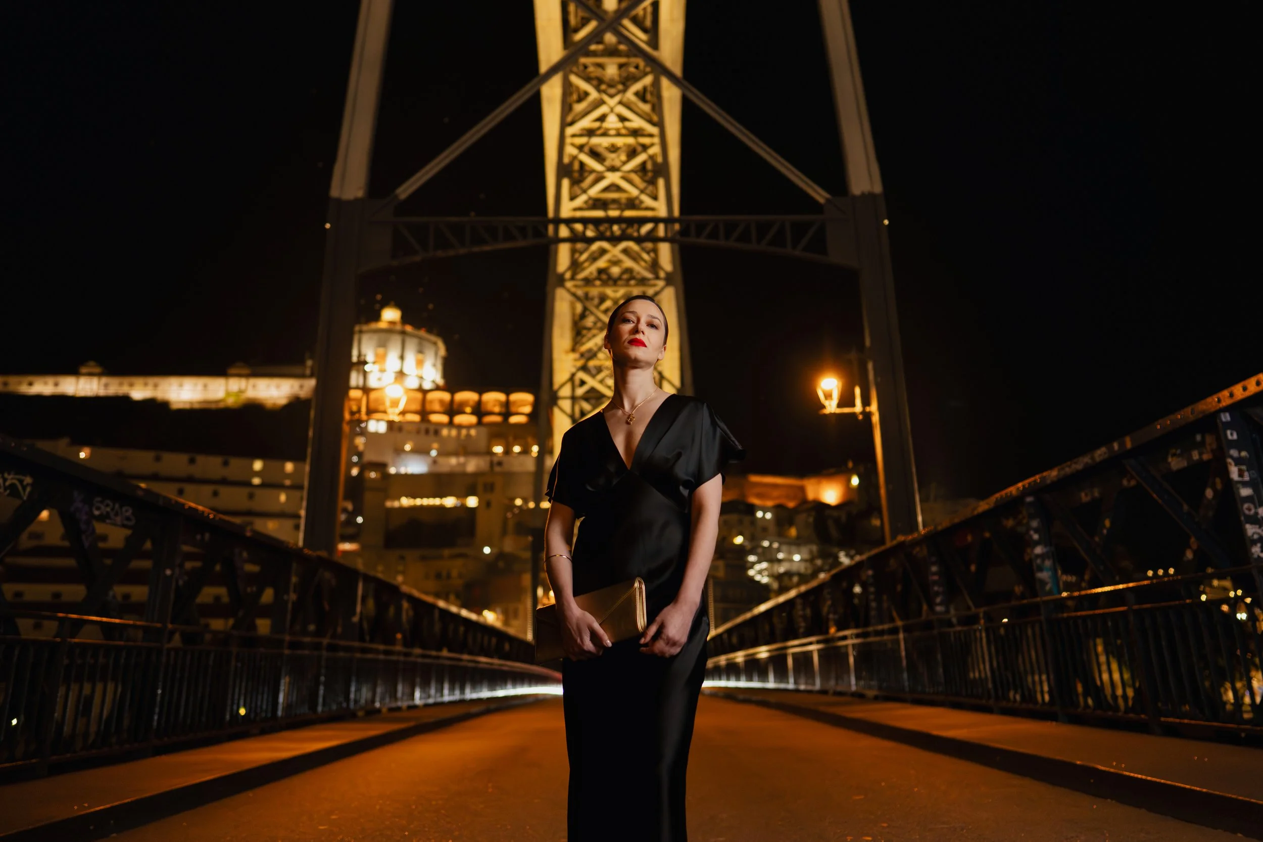 Femme en robe noire élégante tenant un porte-documents doré, debout sur un pont suspendu la nuit avec la tour Eiffel illuminée en arrière-plan.