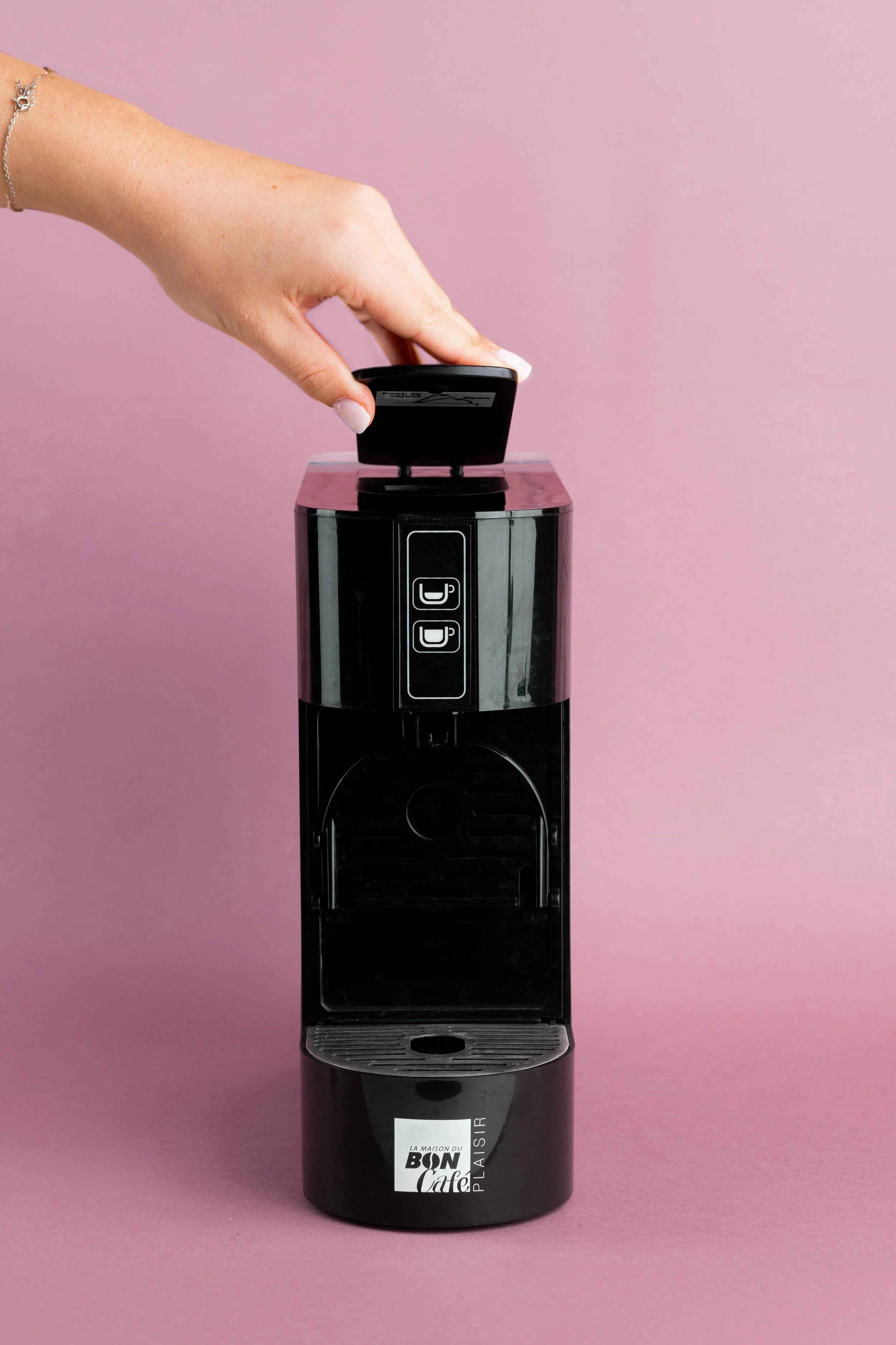 Une main insère une capsule de café dans une machine à café noire contre un fond rose.
