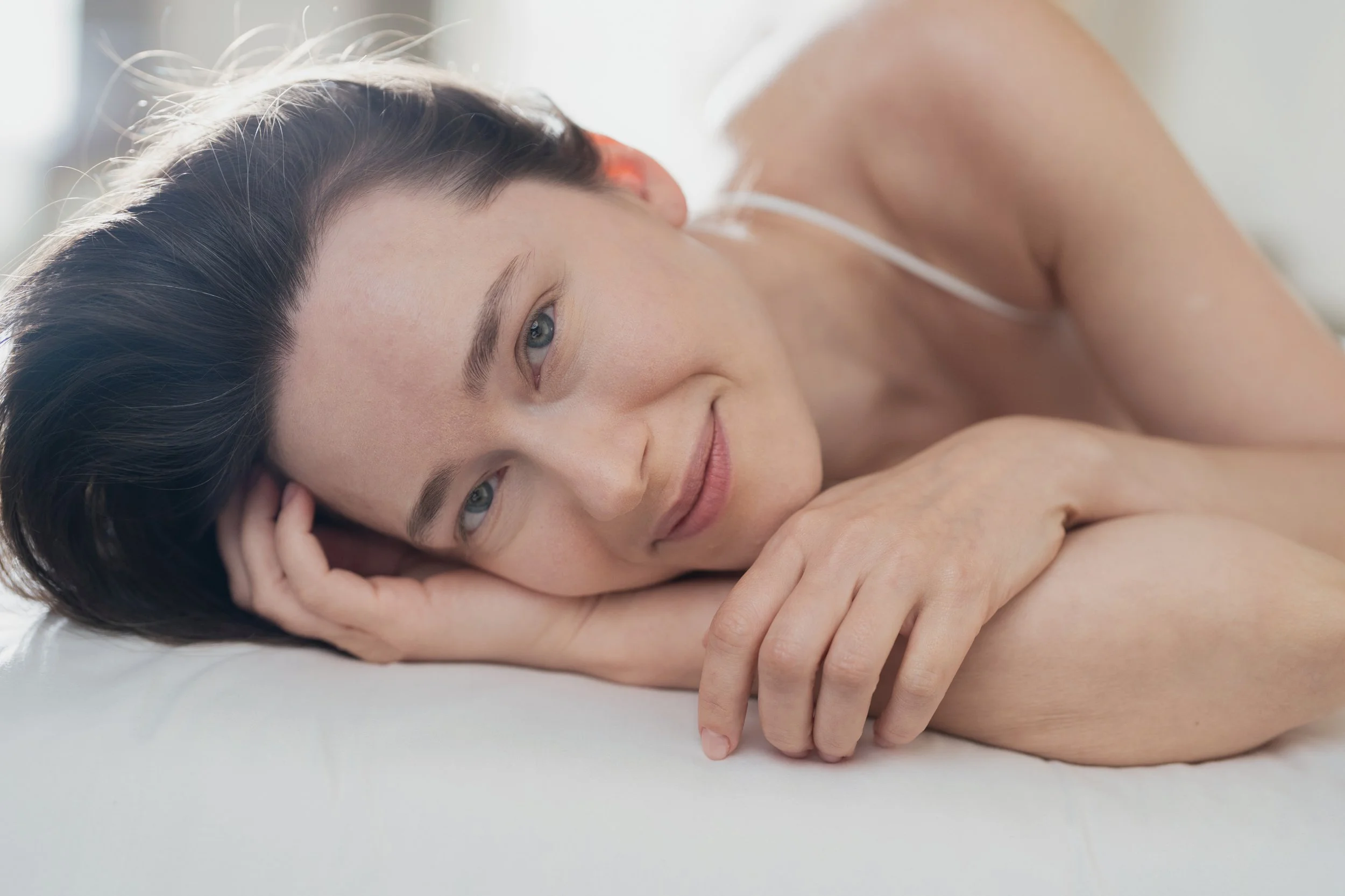 Femme rêveuse allongée sur un lit, souriant doucement avec la tête reposant sur le bras, portant un haut blanc, dans un environnement lumineux.