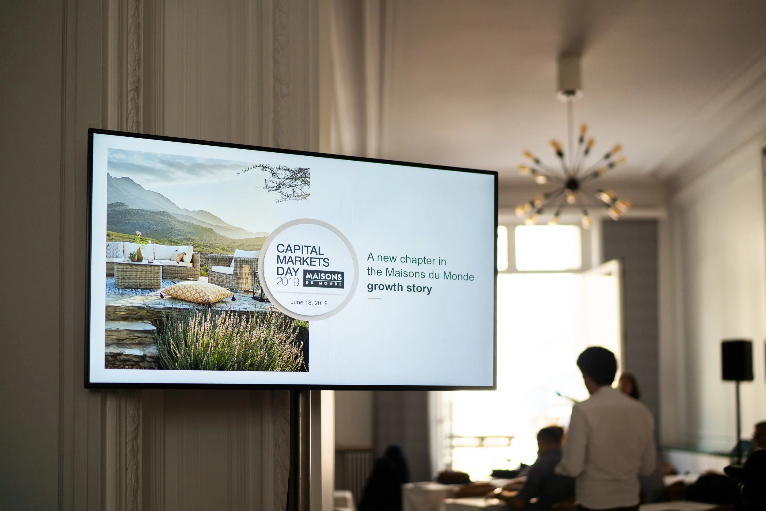 Une télévision affiche une présentation sur la Journée du Marché des Capitaux 2019, avec une photo d'un mobilier d'extérieur et un texte en anglais et français.