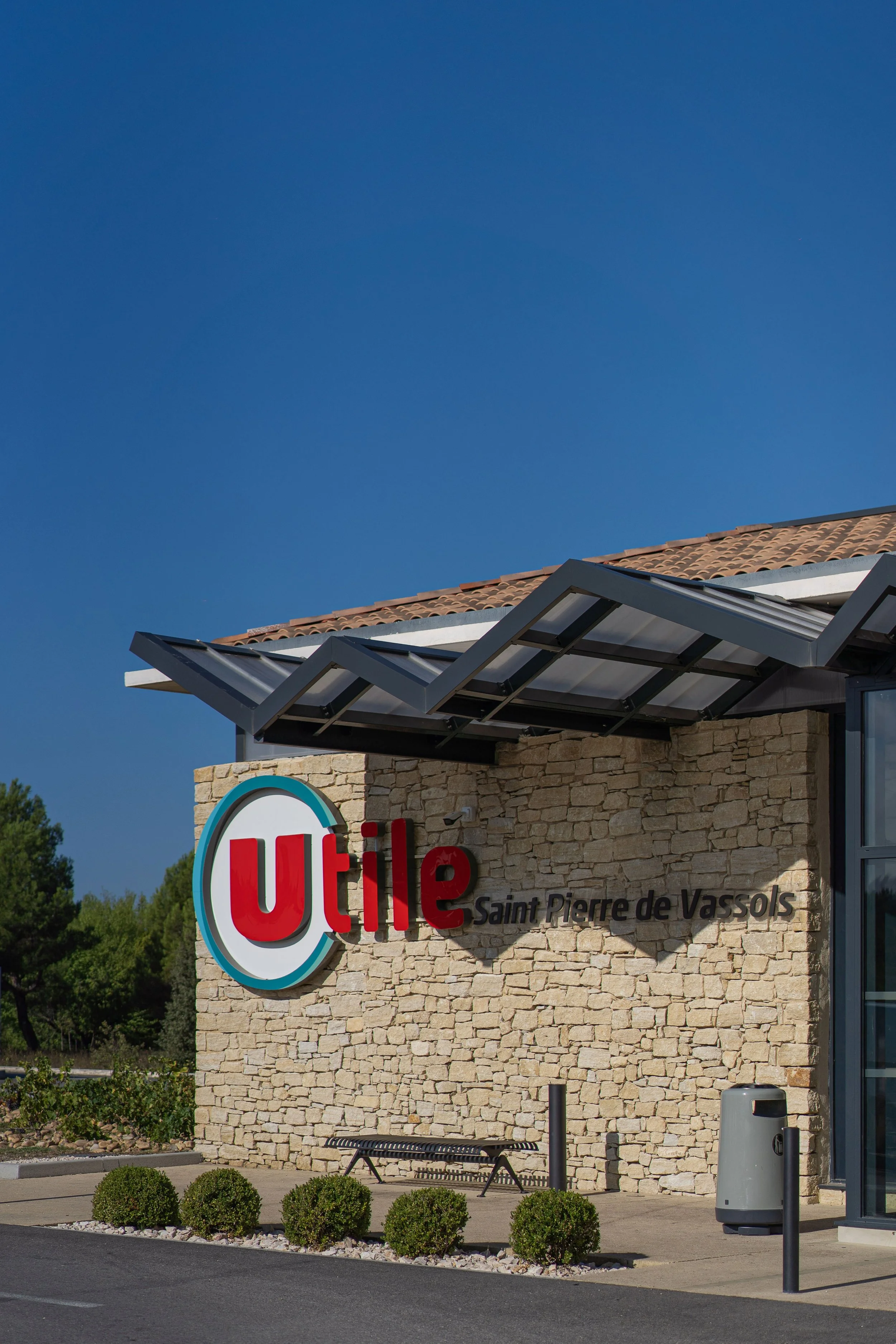 Façade d'un bâtiment avec un panneau indiquant 'Utile Saint Pierre de Vassols', un banc, des arbustes et un poubelle, sous un ciel bleu.