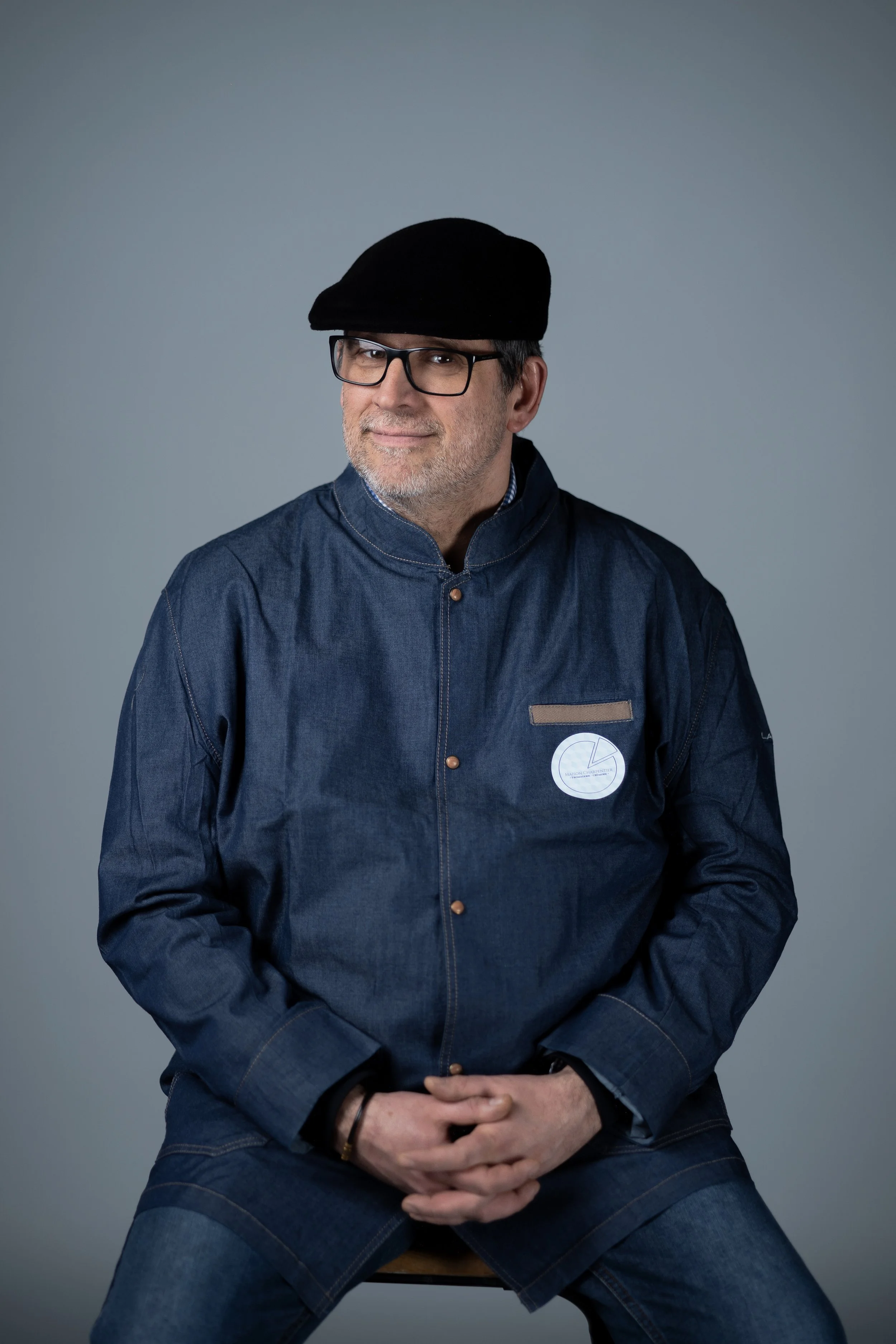 Homme âgé portant une veste en jean bleu, une casquette noire, des lunettes et un bracelet, assis sur une chaise, avec un fond gris