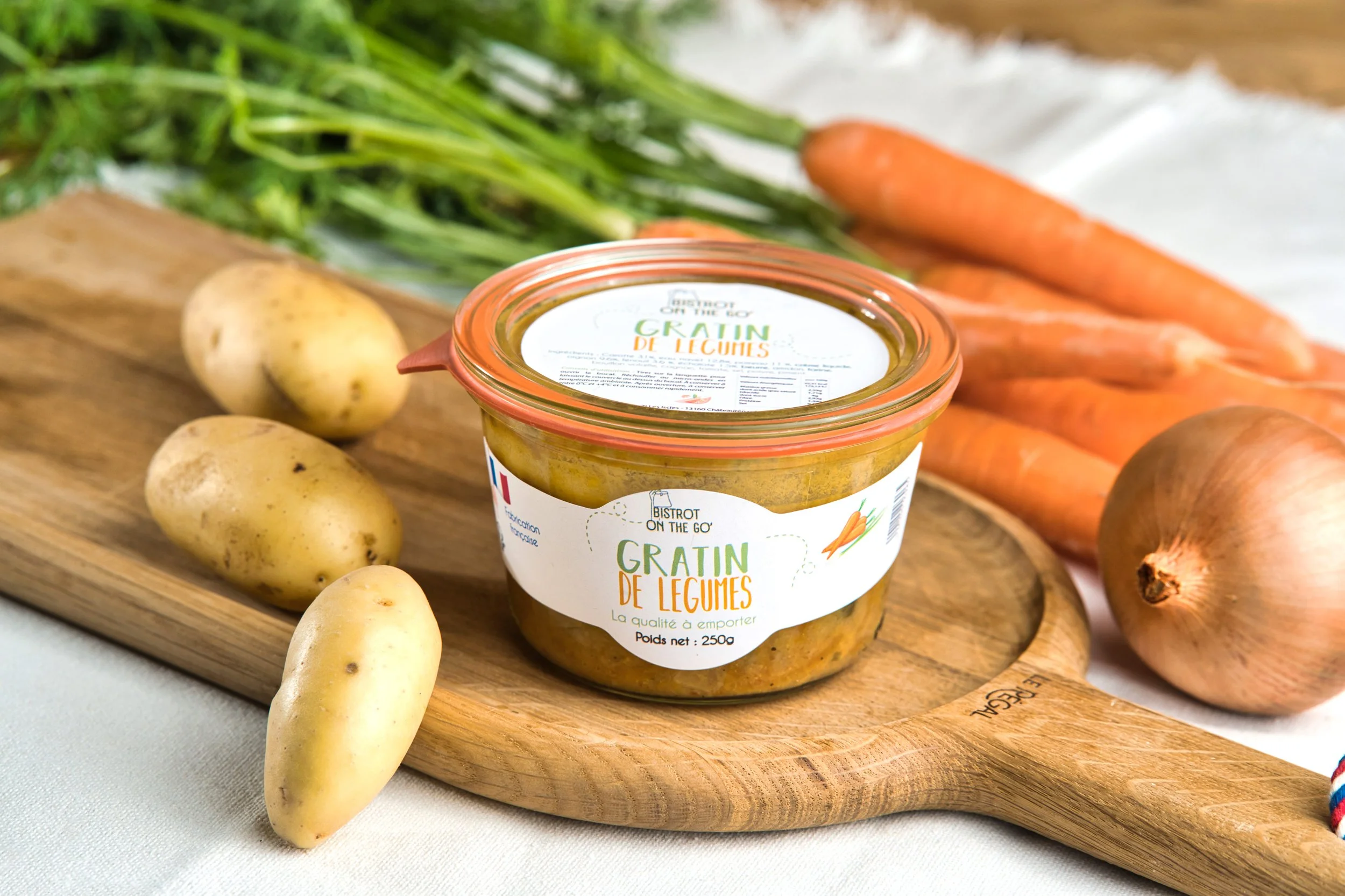 Pot de gratin de légumes avec des pommes de terre, des carottes et des oignons autour, sur une planche en bois.