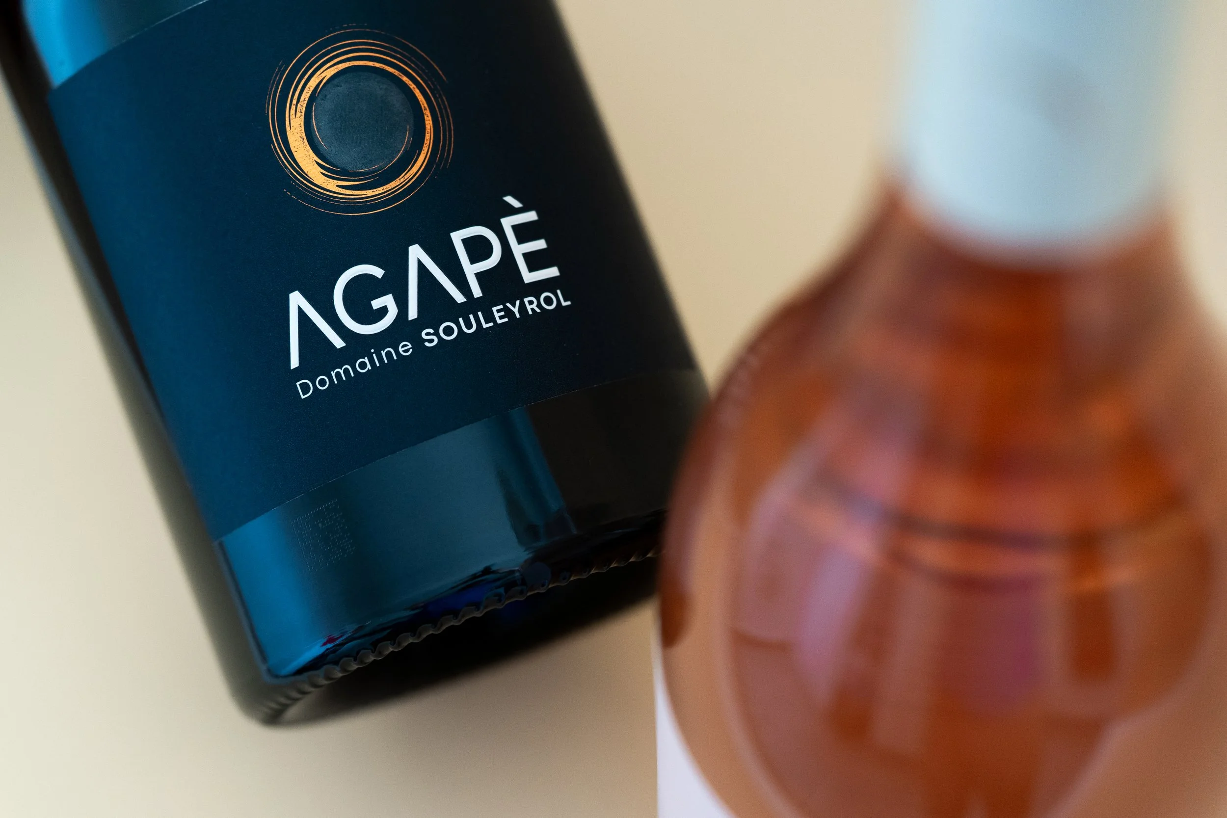 Bouteille de vin rouge Domaine Souleyrol Agapé avec un label noir et un cercle doré au sommet, en partie visible, à côté d'une bouteille de couleur orange avec un bouchon blanc.