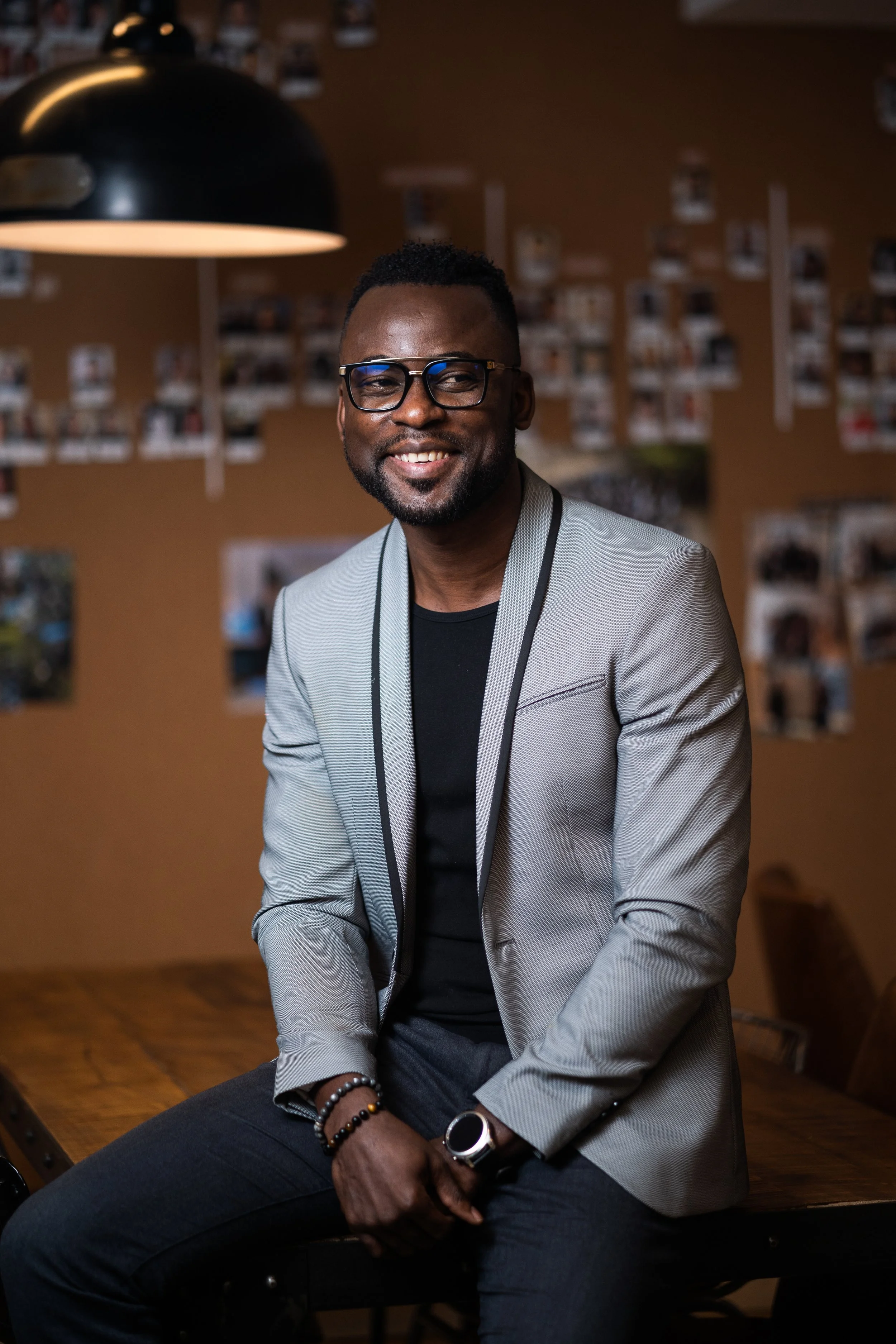 Un homme souriant, portant des lunettes, une veste grise et une montre, assis sur une table en bois dans une pièce décorée avec des photos au mur.