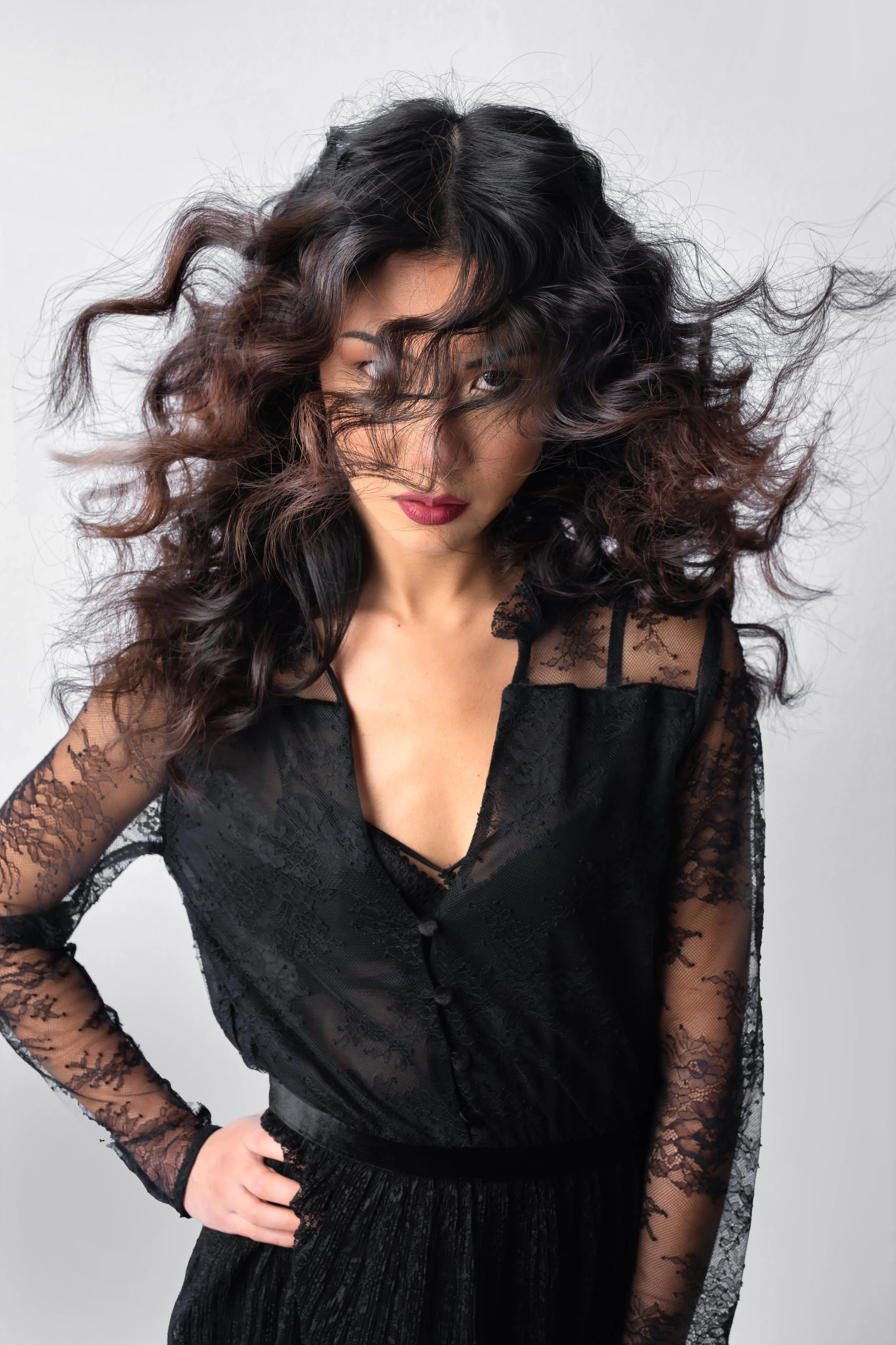 Femme avec cheveux bouclés, portant une robe en dentelle noire, pose avec une main sur la taille, fond gris.