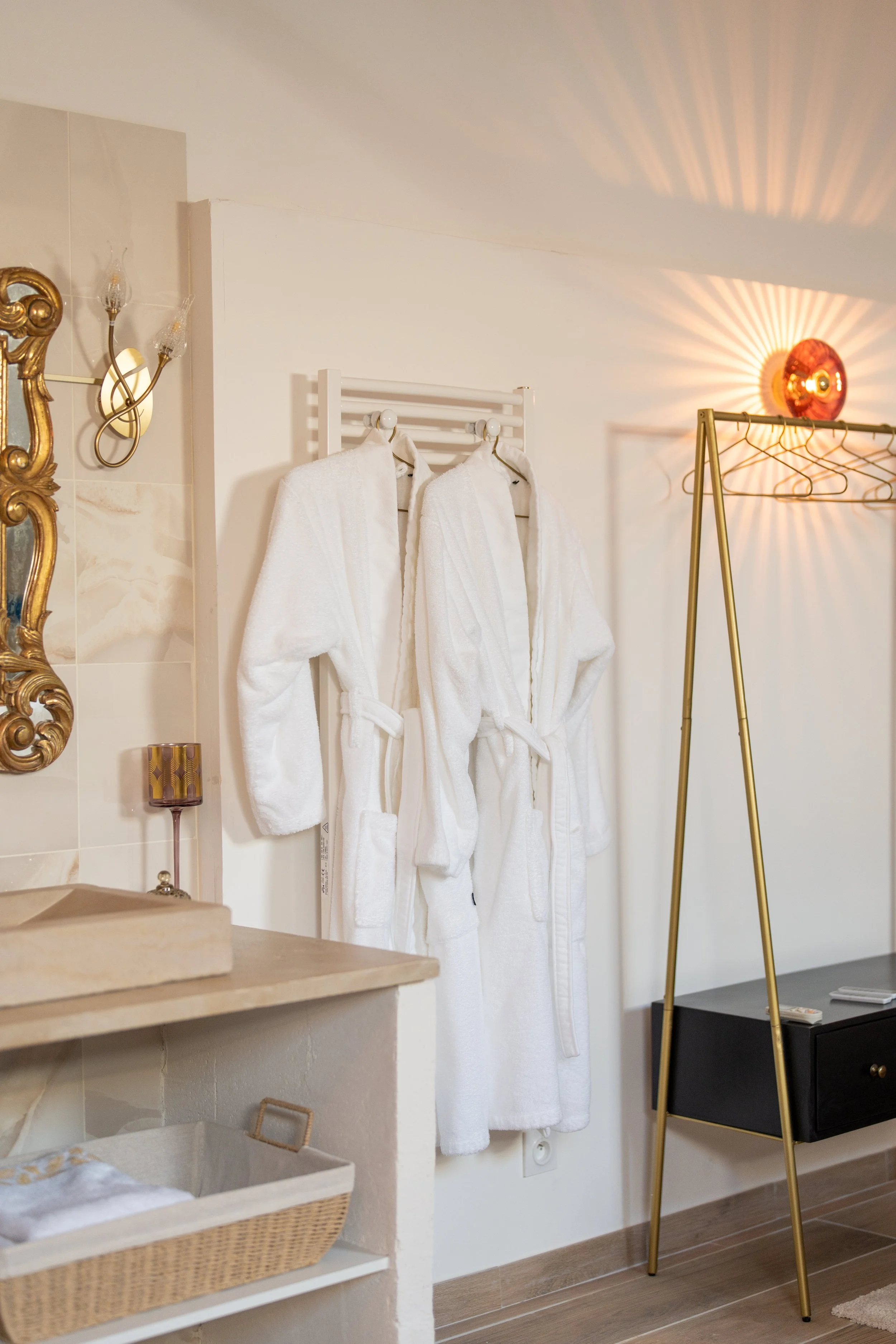 Deux peignoirs blancs suspendus dans une salle de bain ou chambre, avec un miroir doré, une lampe murale et une lampe rouge projetant de la lumière sur le mur, un porte-vêtements en métal doré, un meuble noir, et une salle de bain partielle visible.