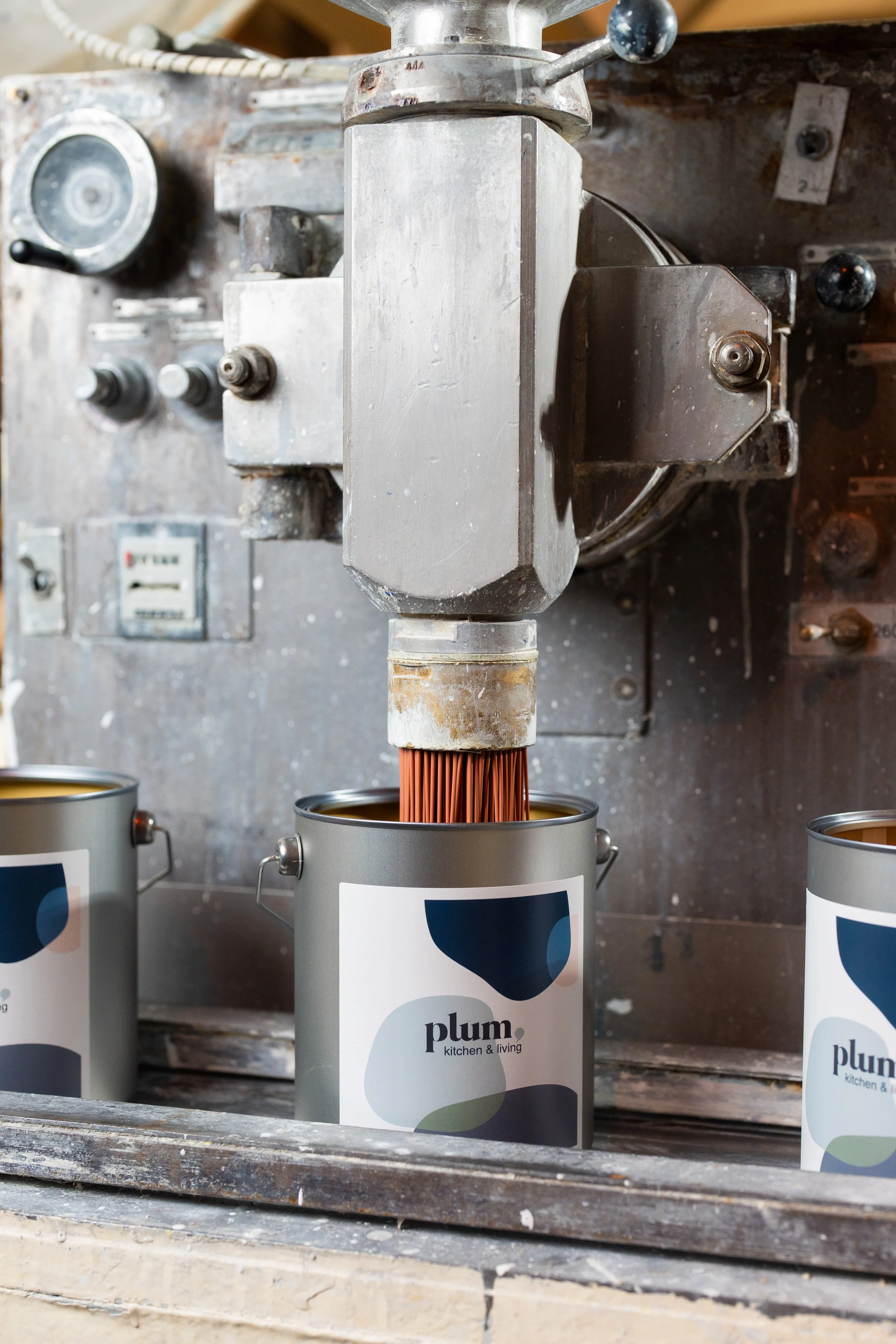 Une usine ou une machine de fabrication de peinture, avec des bidons de peinture étiquetés 'plum kitchen & living' sur un plan de travail en bois.