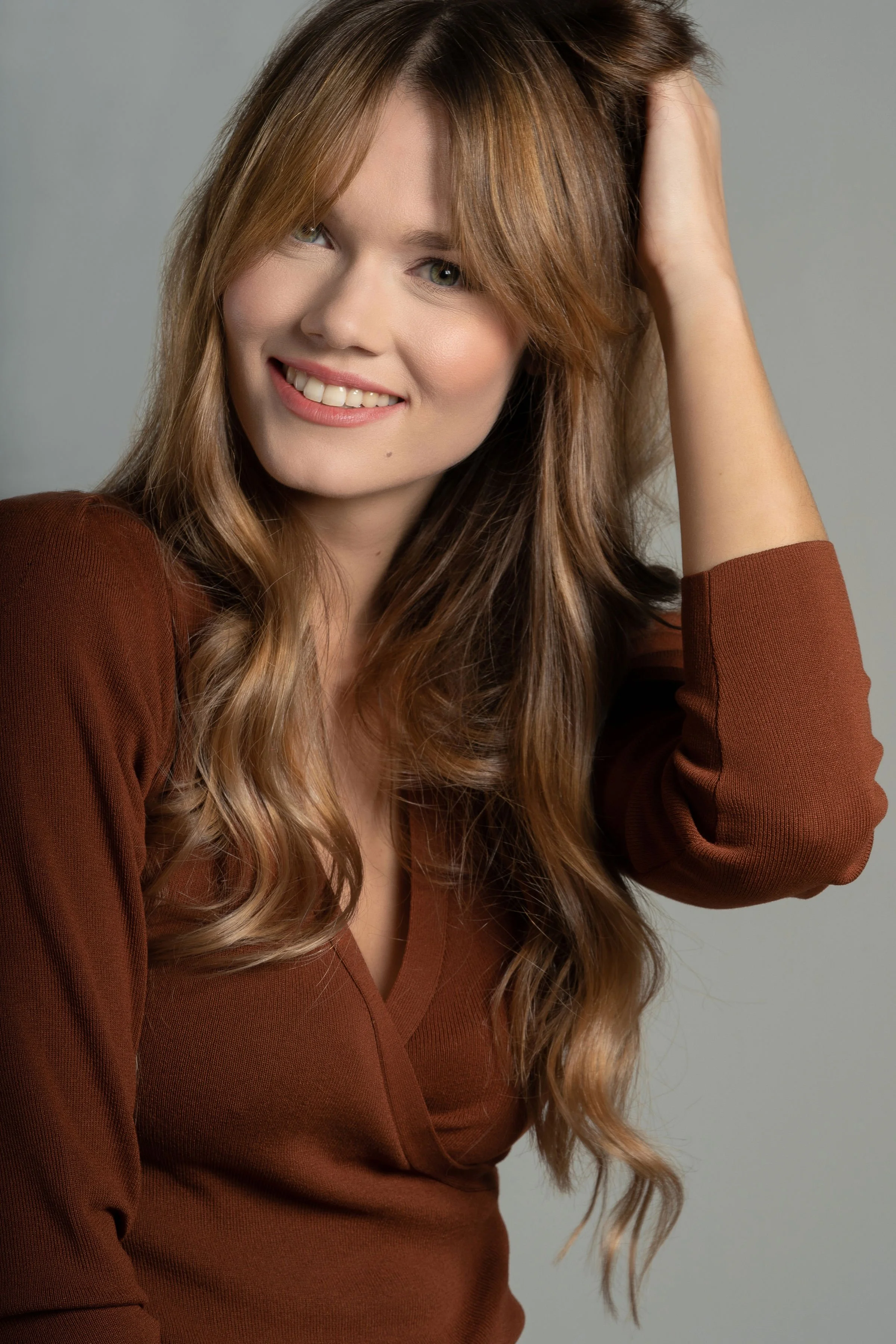 Jeune femme aux cheveux longs et bruns flottants, souriant, portant un haut marron avec un fond gris clair.