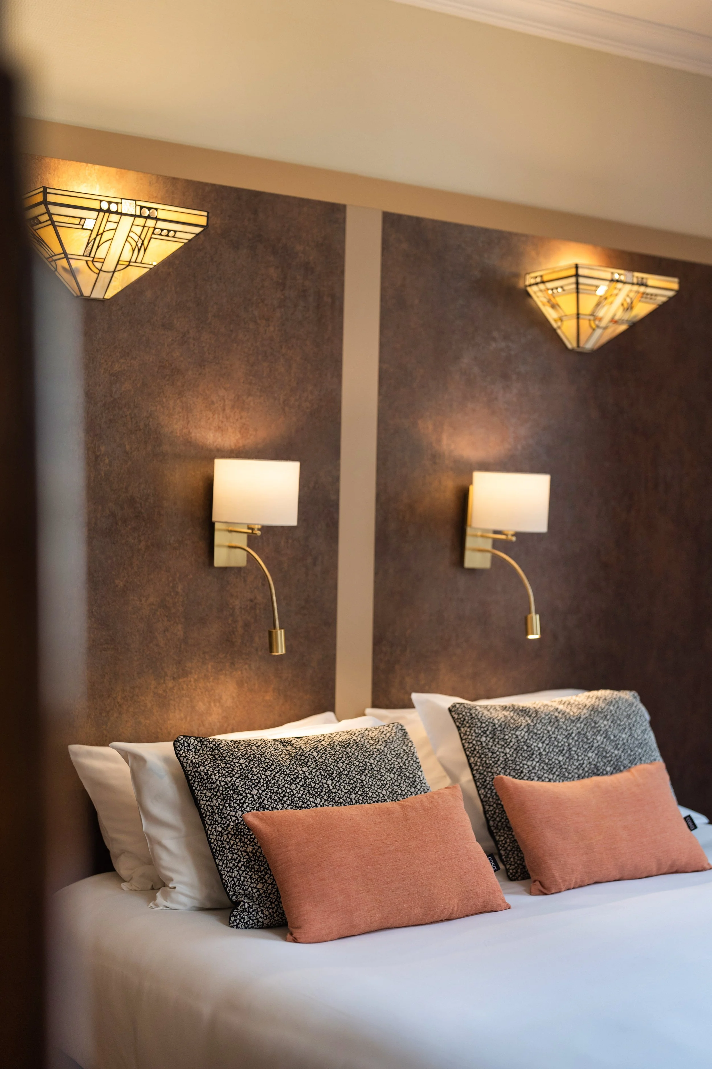 Un lit dans une chambre d'hôtel avec deux coussins noirs et deux coussins roses, deux appliques murales et deux lampes de chevet, décor modernes et élégants.