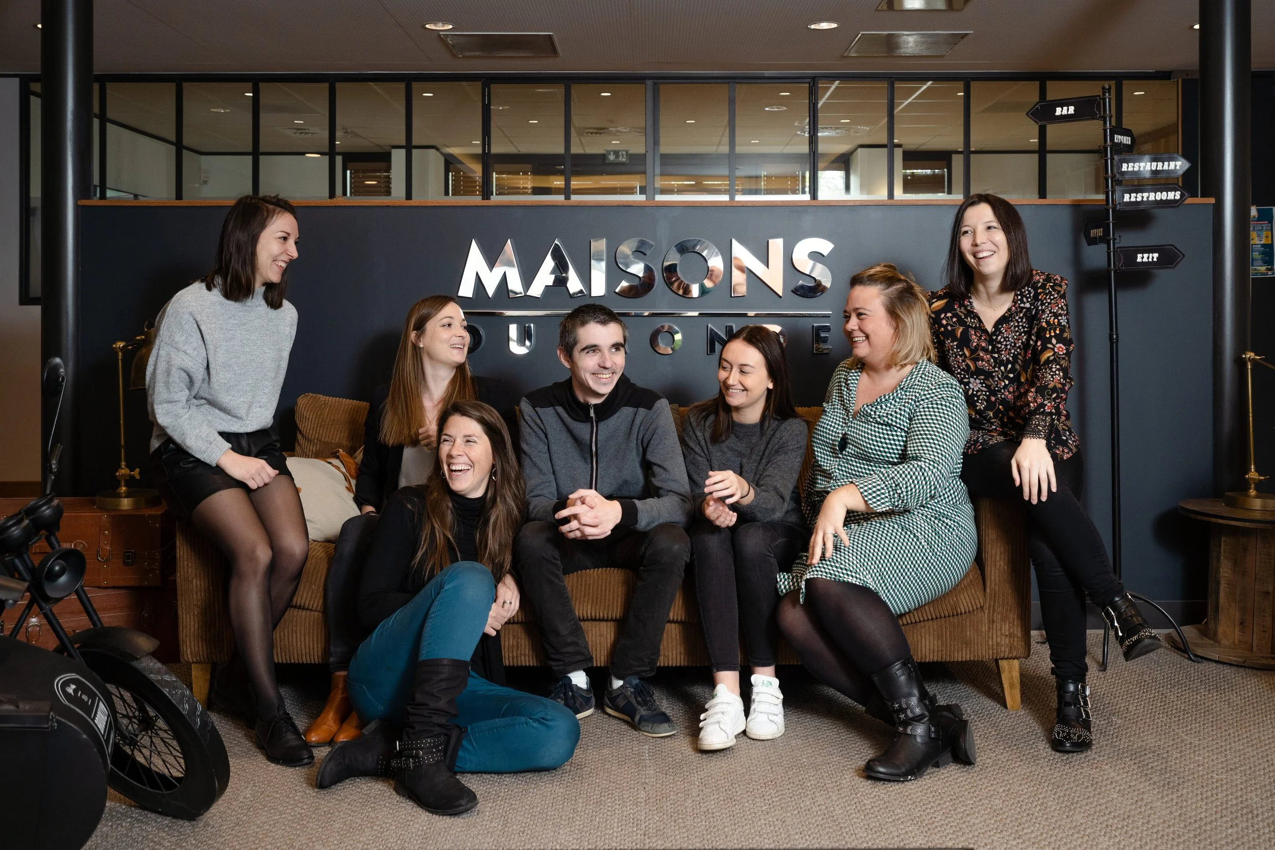 Groupe de sept personnes souriantes assises sur un canapé en intérieur, avec un panneau en arrière-plan indiquant "Maisons du Monde" et un signe directionnel pointant vers bar, restaurant, toilettes et sortie.
