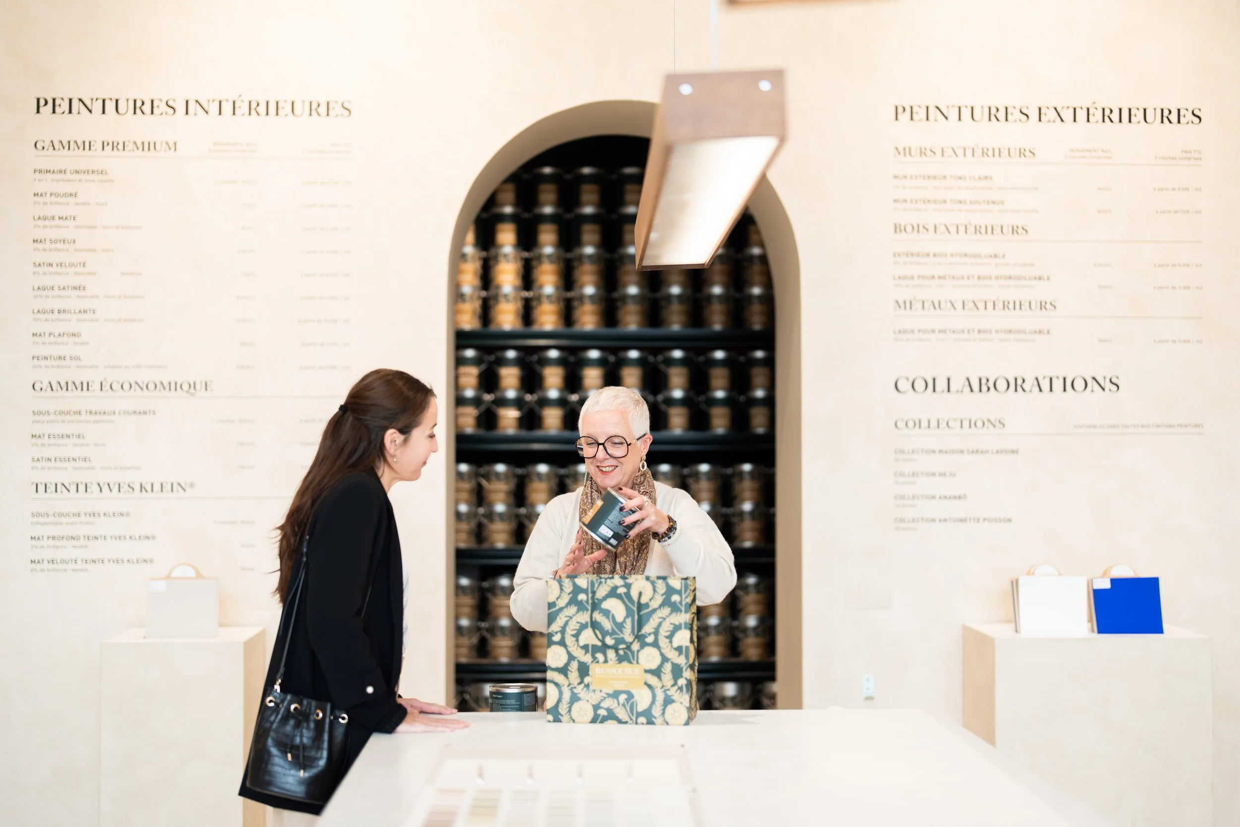 Une femme âgée avec des cheveux gris, portant des lunettes, échange un produit avec une femme plus jeune dans un magasin, avec des étagères d'articles en arrière-plan.