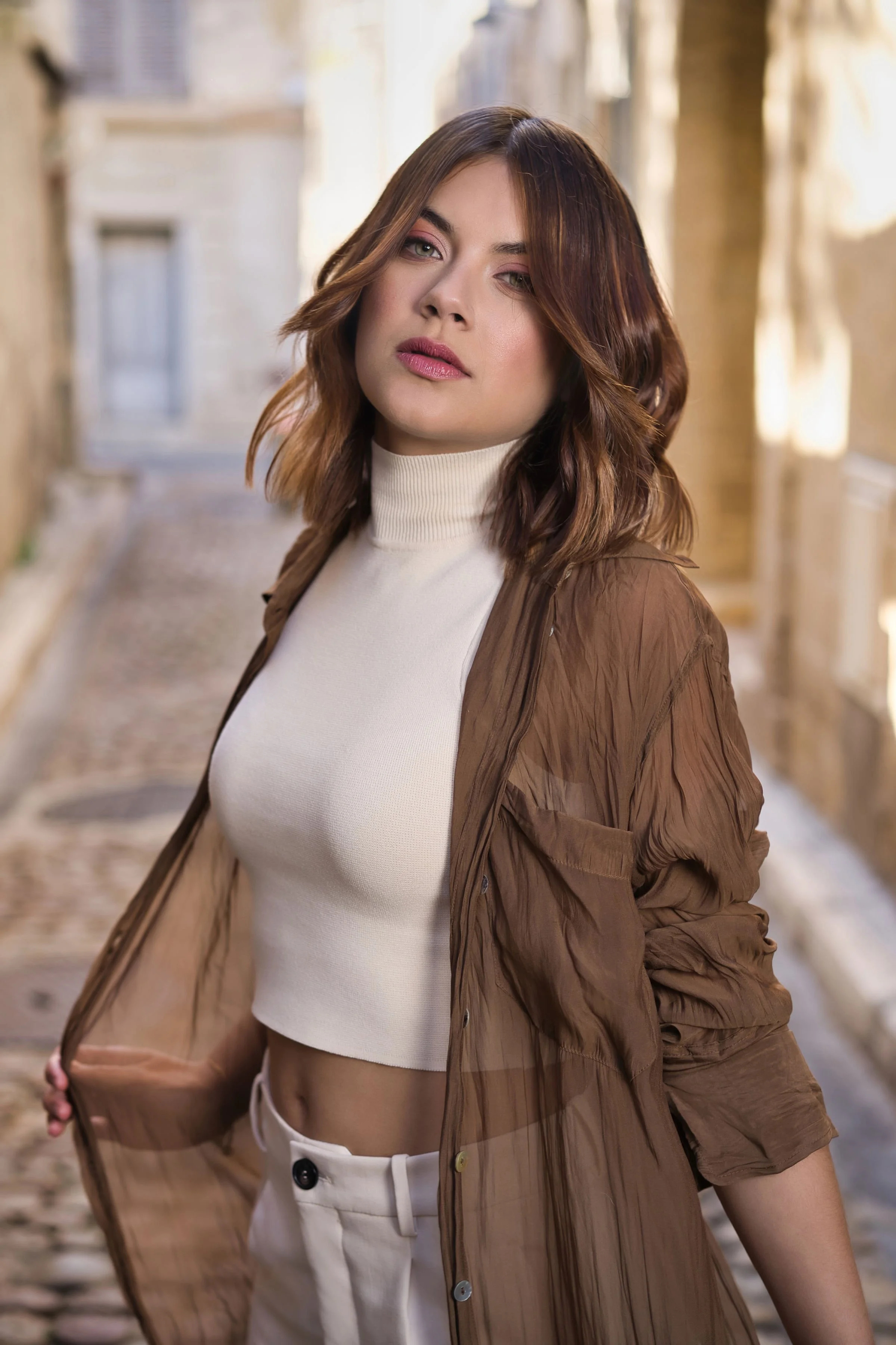 Une femme avec des cheveux bruns et ondulés, portant un pull blanc, une veste en tissu transparent marron et un pantalon blanc, se tient dans une ruelle pavée avec des bâtiments anciens en arrière-plan.