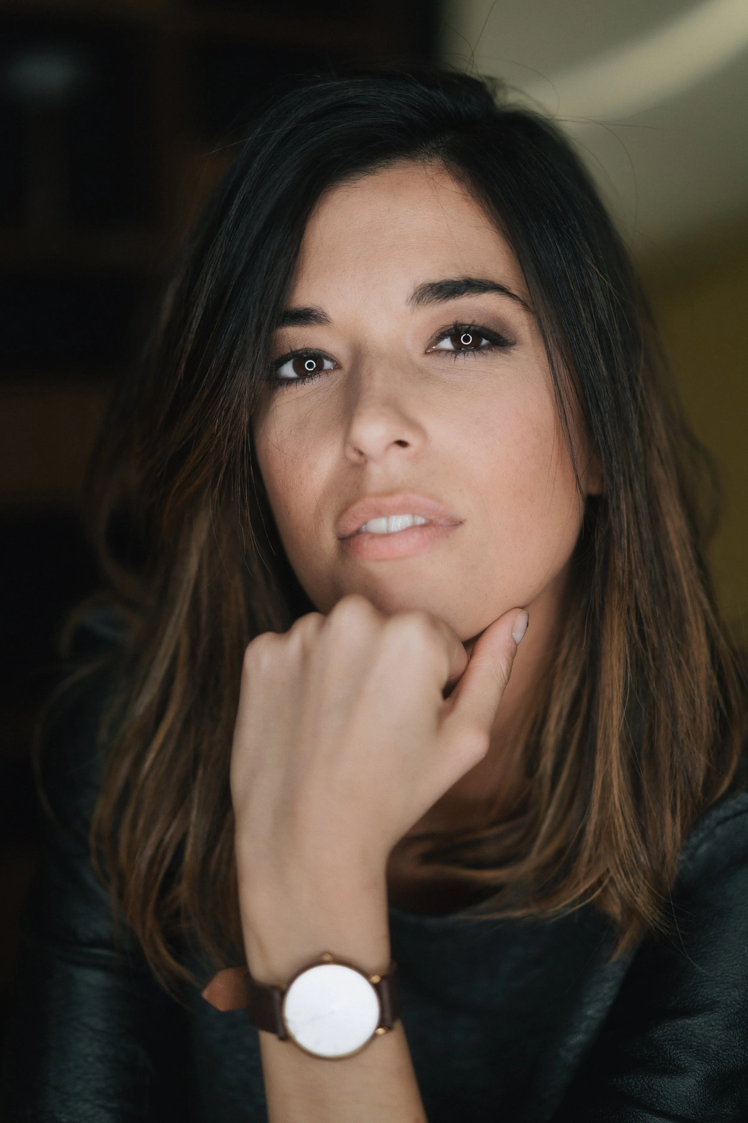 Jeune femme aux cheveux bruns, regardant l'objectif, avec la main sous le menton, portant une montre blanche et un bracelet, contre un fond flou.