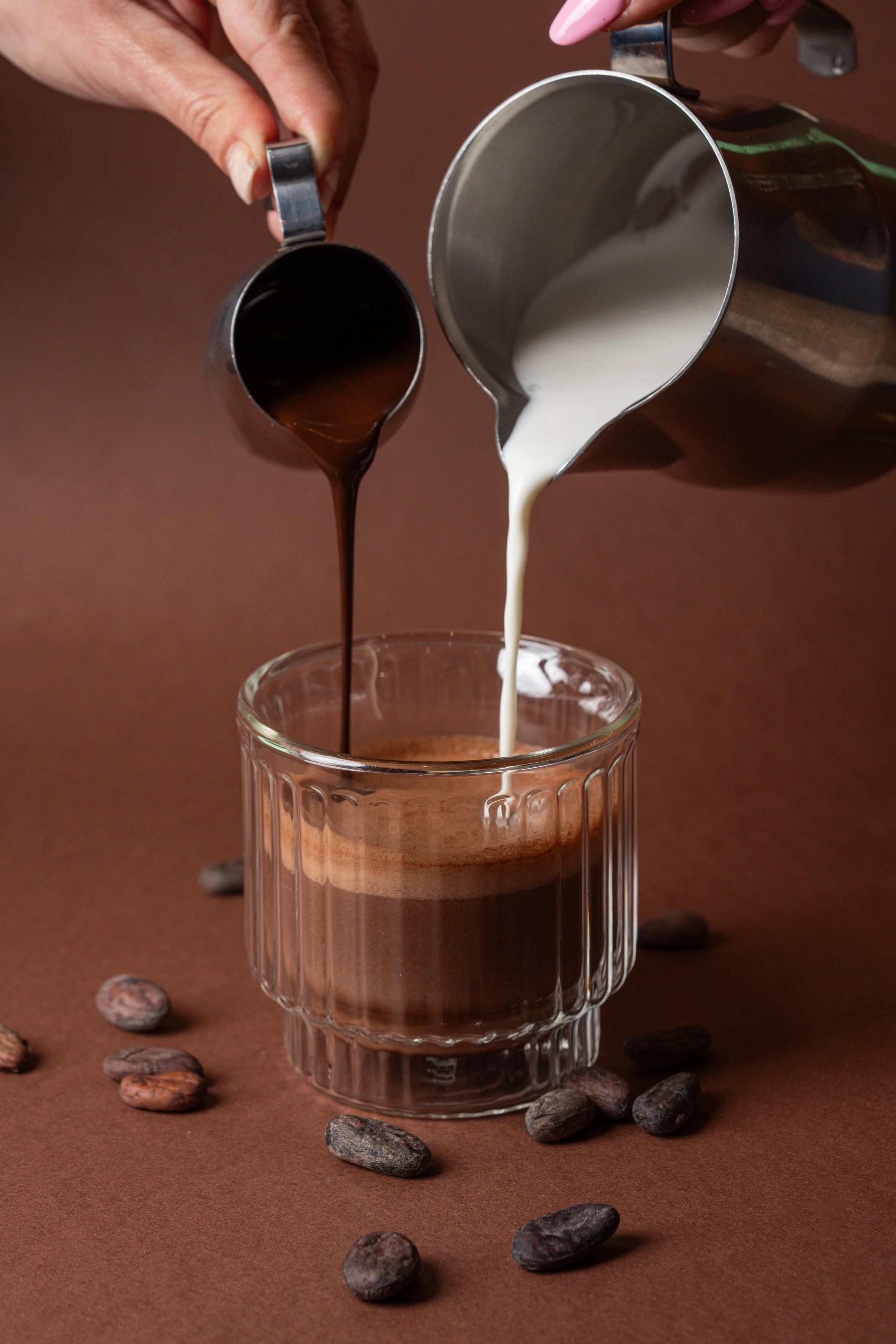 Préparation d'un café avec du lait versé dans une tasse en verre, entourée de grains de café sur une surface marron.