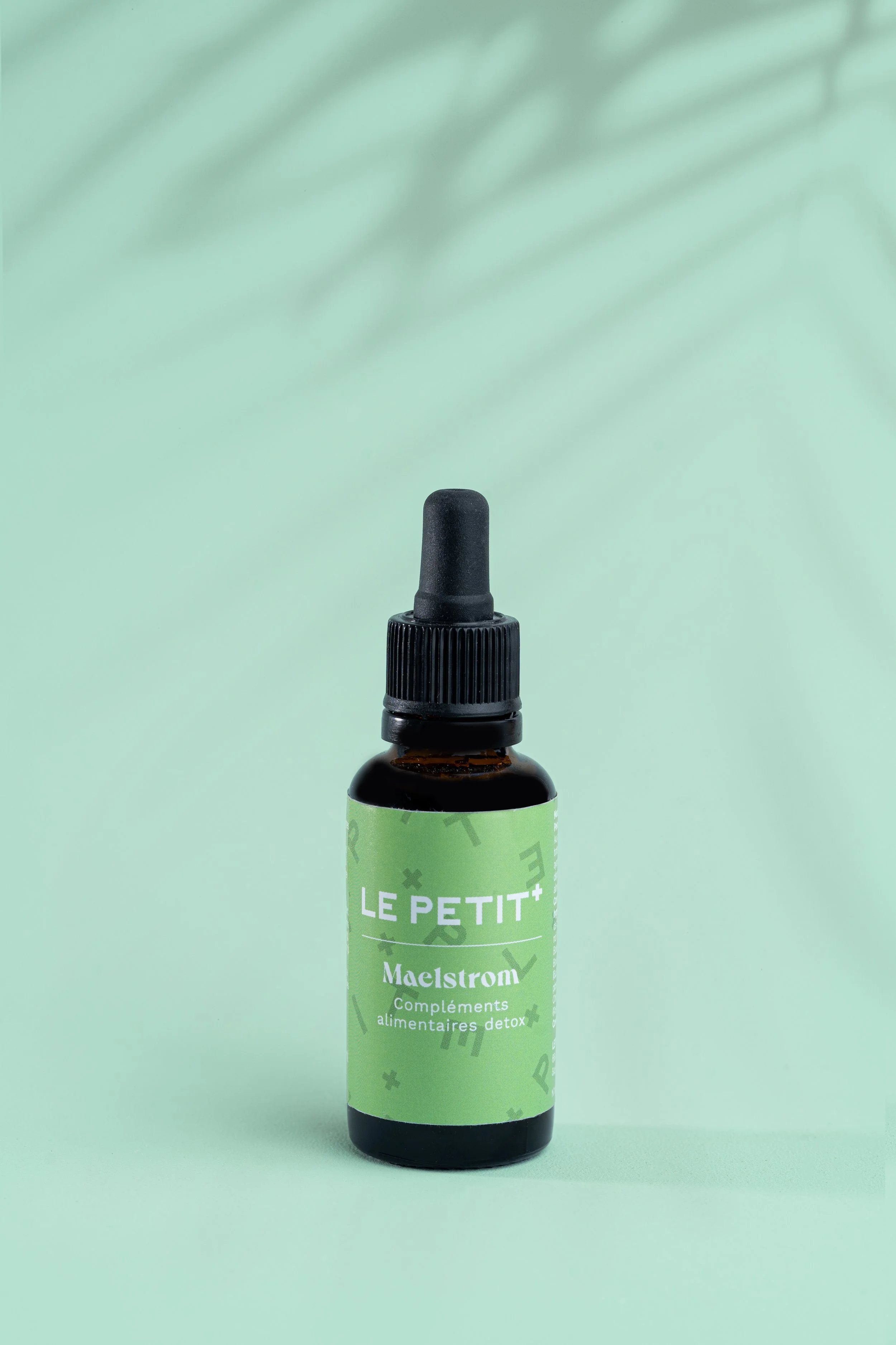 Un flacon brown avec un bouchon compte-gouttes noir, étiqueté 'LE PETIT+', contenant des compléments alimentaires détox 'Maelstrom', sur un fond vert clair avec ombres de feuilles