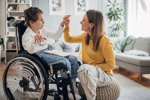 A young girl in a wheelchair giving a high five to a woman in a yellow sweater indoors with a sofa and plants in the background.