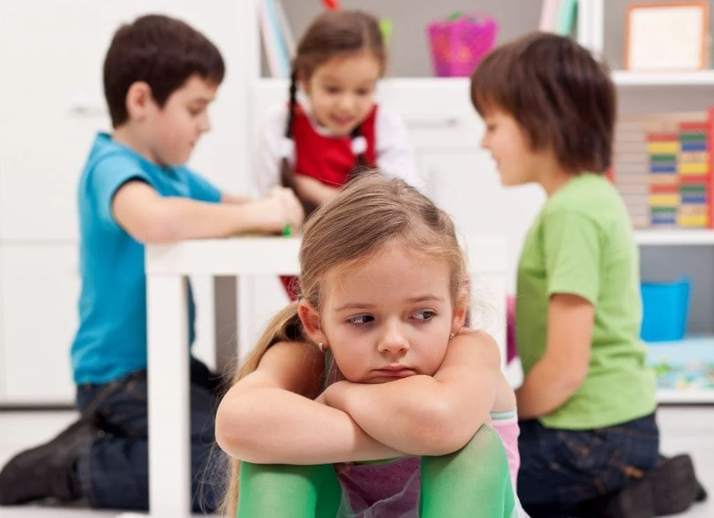 A young girl looks sad and bored, sitting on the floor with her arms resting on her knees, as three children play and interact happily behind her.