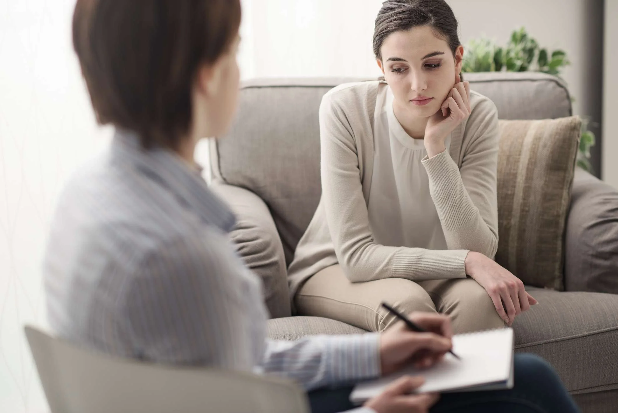A woman sitting on a couch during a therapy session, with a therapist taking notes.