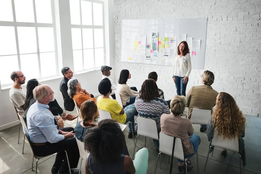 A woman giving a presentation to a diverse group of people in a bright, modern room with large windows and white brick walls.