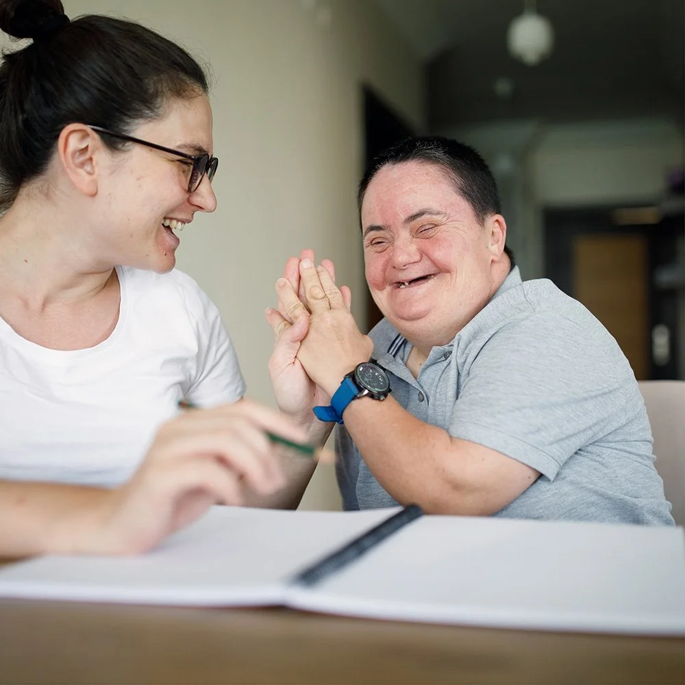 A woman and a man are smiling and holding hands in a gesture of friendship or support inside a home or office with a blurred background.