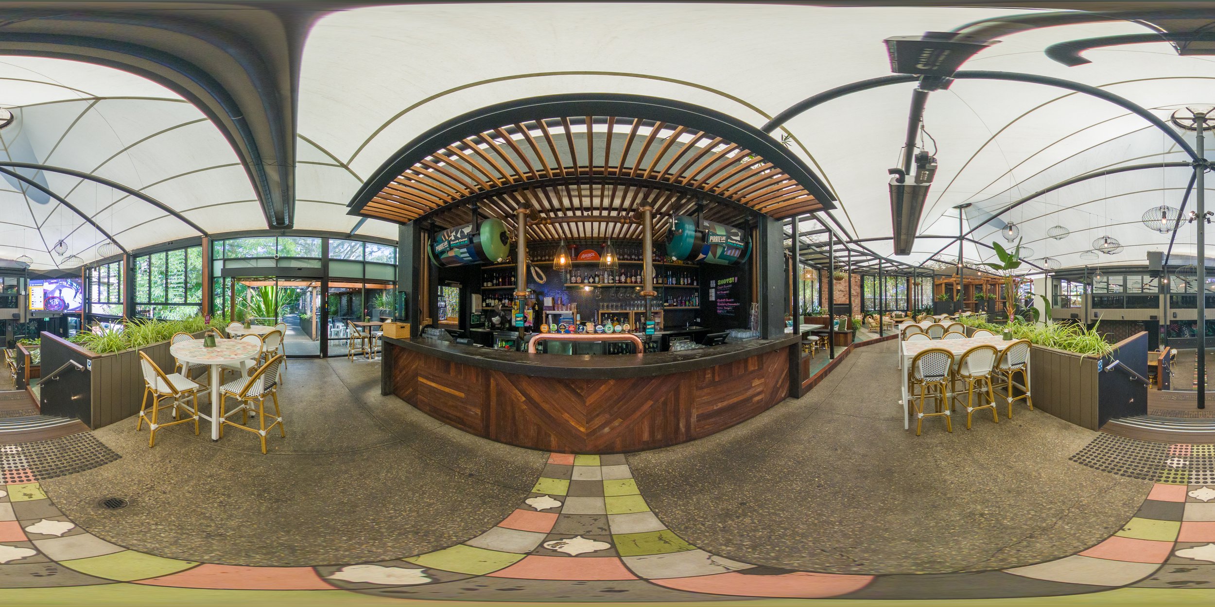 Interior of a bright, modern restaurant with a wooden bar in the center, surrounded by tables and chairs, and large windows letting in natural light.