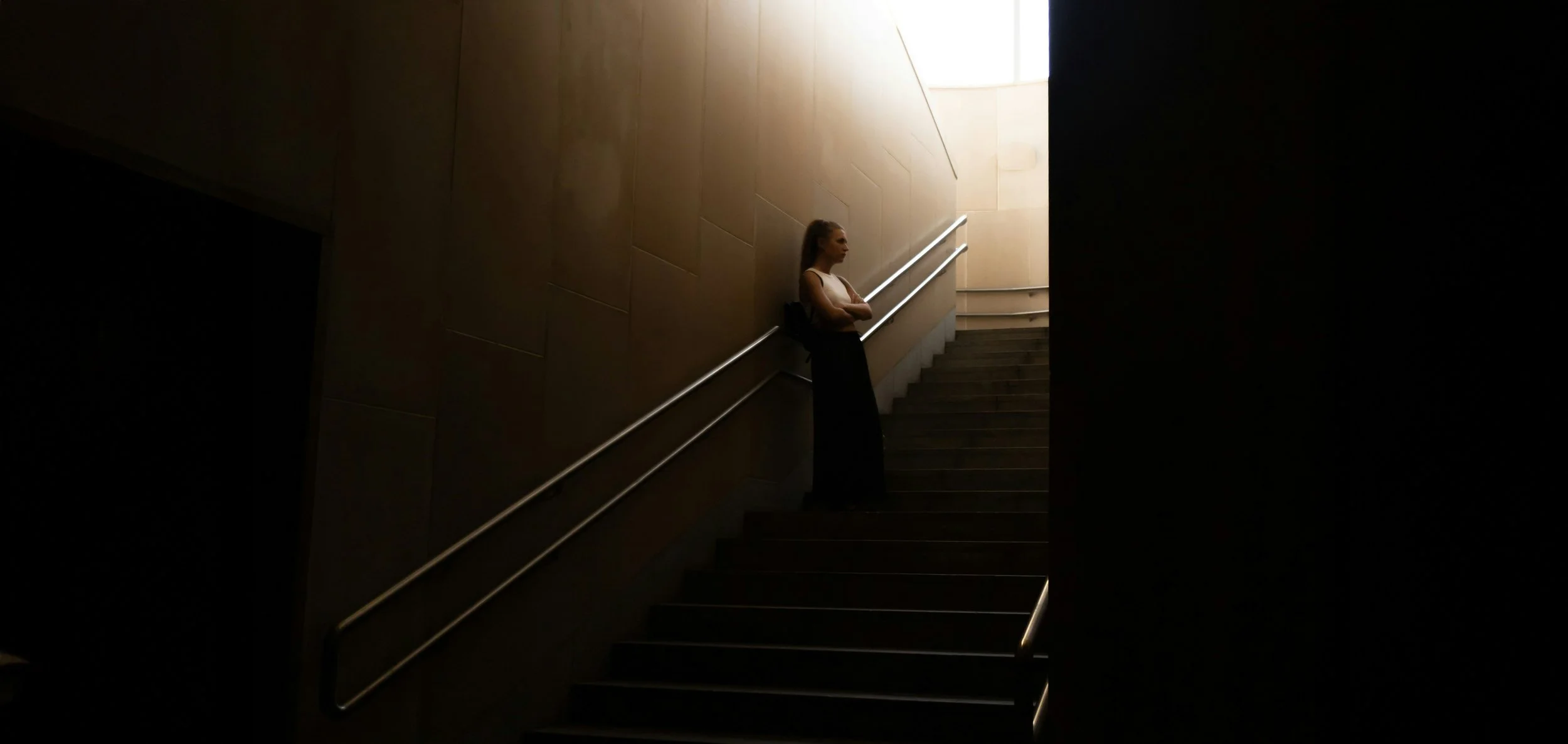 A woman standing on a staircase, leaning against the wall, with her arms crossed, in a dimly lit environment near a bright opening.