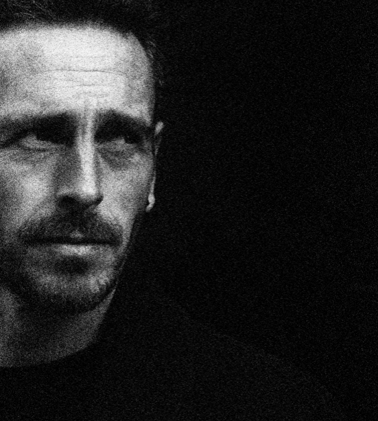 A black-and-white portrait of a man with short dark hair, light facial hair, and a serious expression, against a black background.