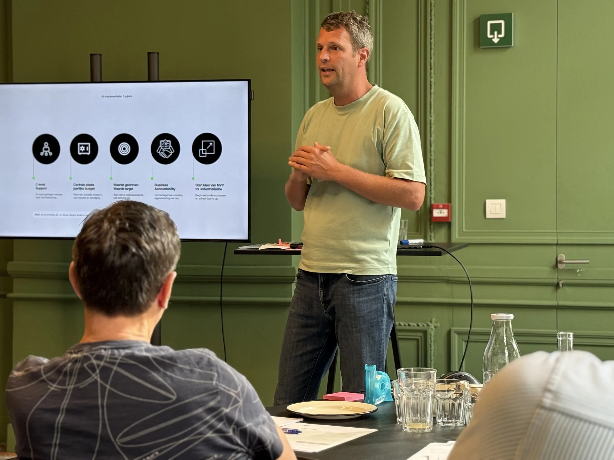 A man giving a presentation in a meeting room with green walls and a large screen displaying slides, while seated attendees listen.