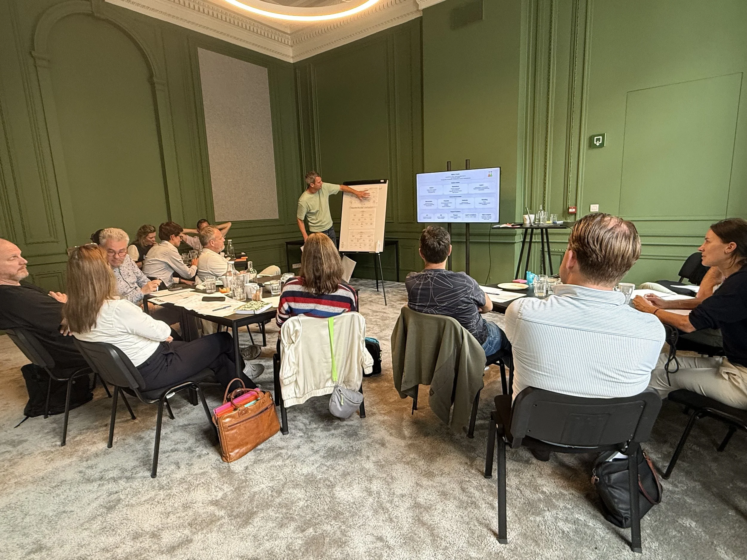 A group of people attending a presentation in a conference room with green walls, listening to a man pointing at a flip chart. A large screen is displaying a flowchart, and there are various table items such as water bottles and notebooks.