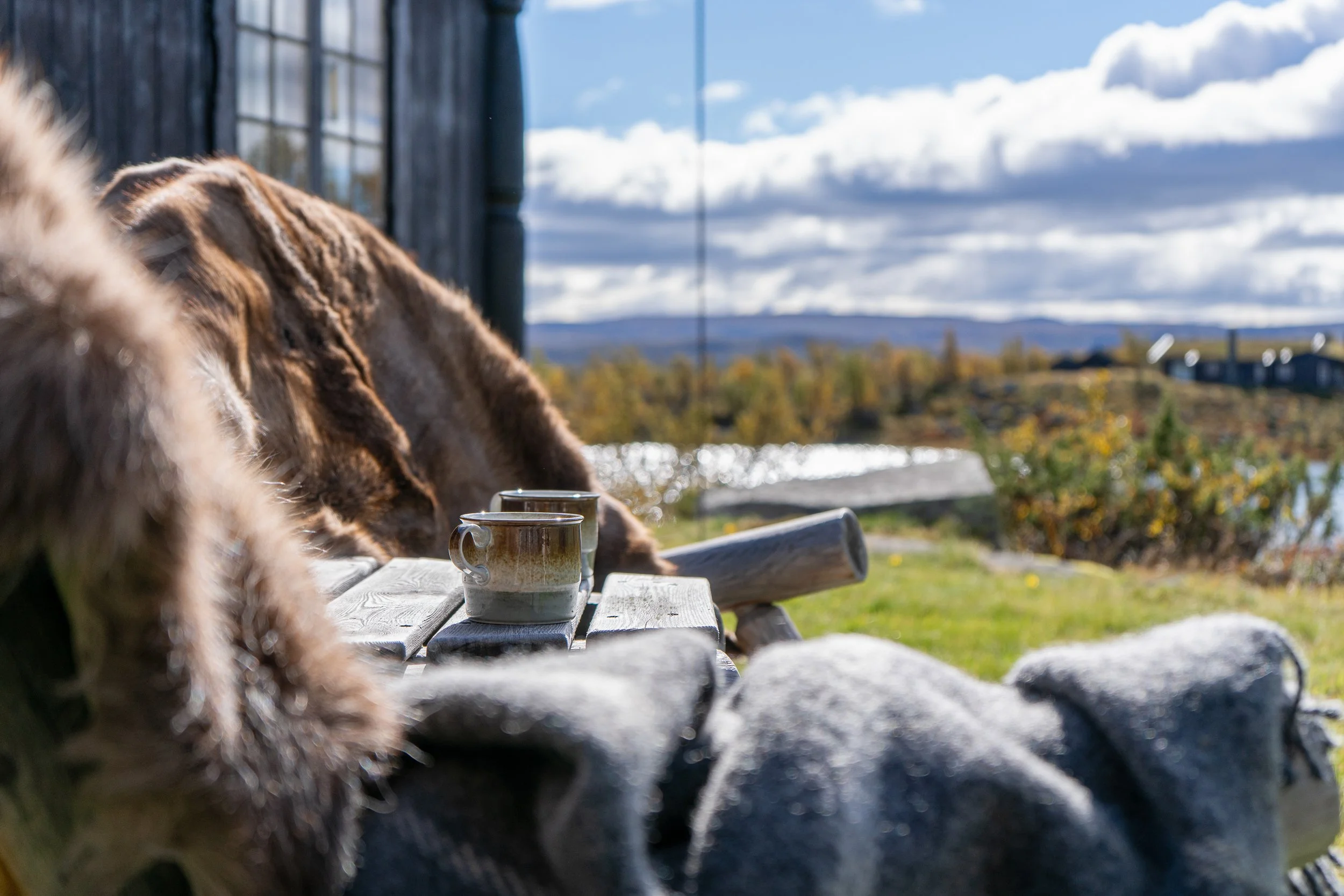 To stoler med saueskinn og tepper ute. Kaffekopper på bordet, og utsikt til vann i bakgrunnen. Foto