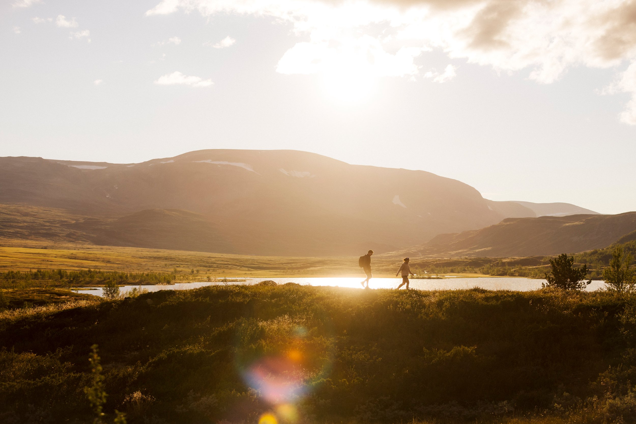 To personer går på fjelltur ved et vann i høstsol.