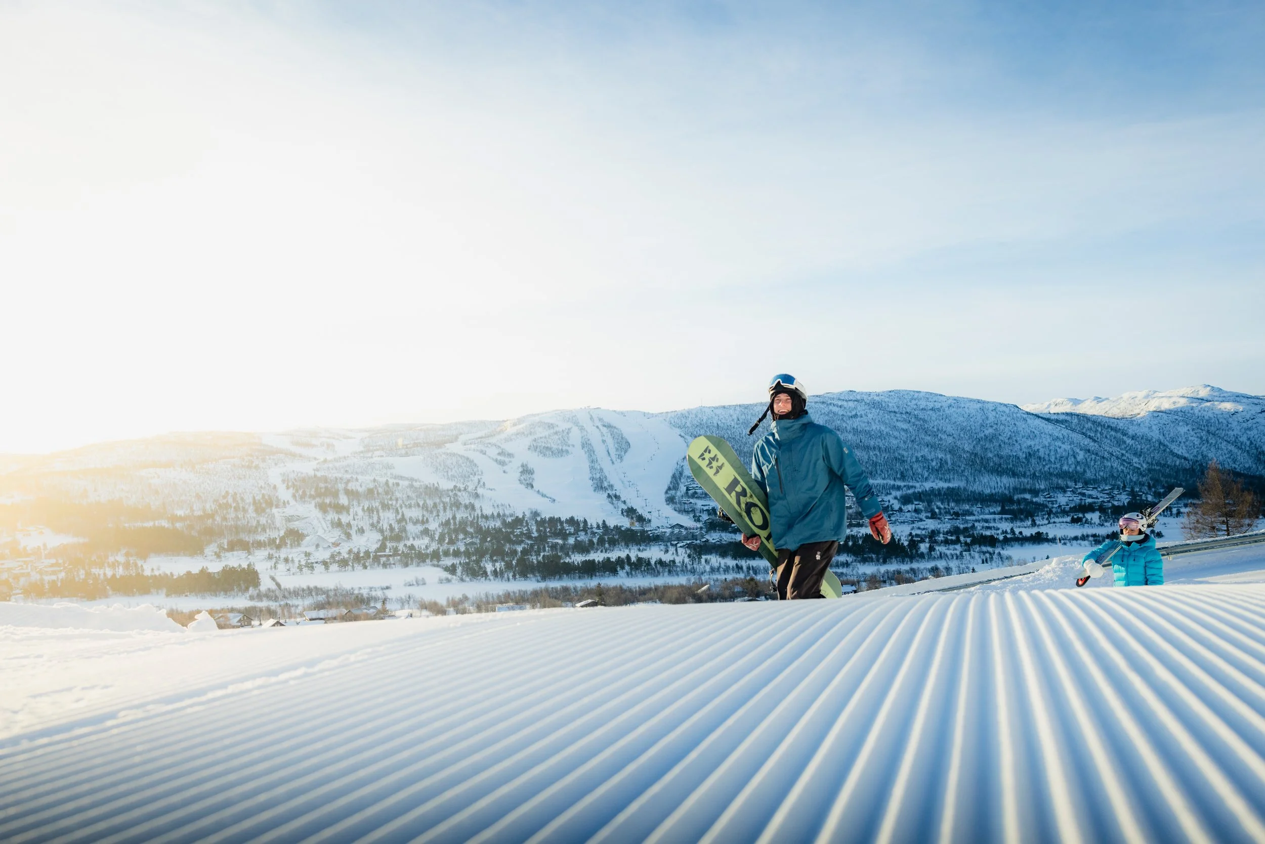 Mann med snowboard og kvinne med ski går oppover skibakken i fint vær.