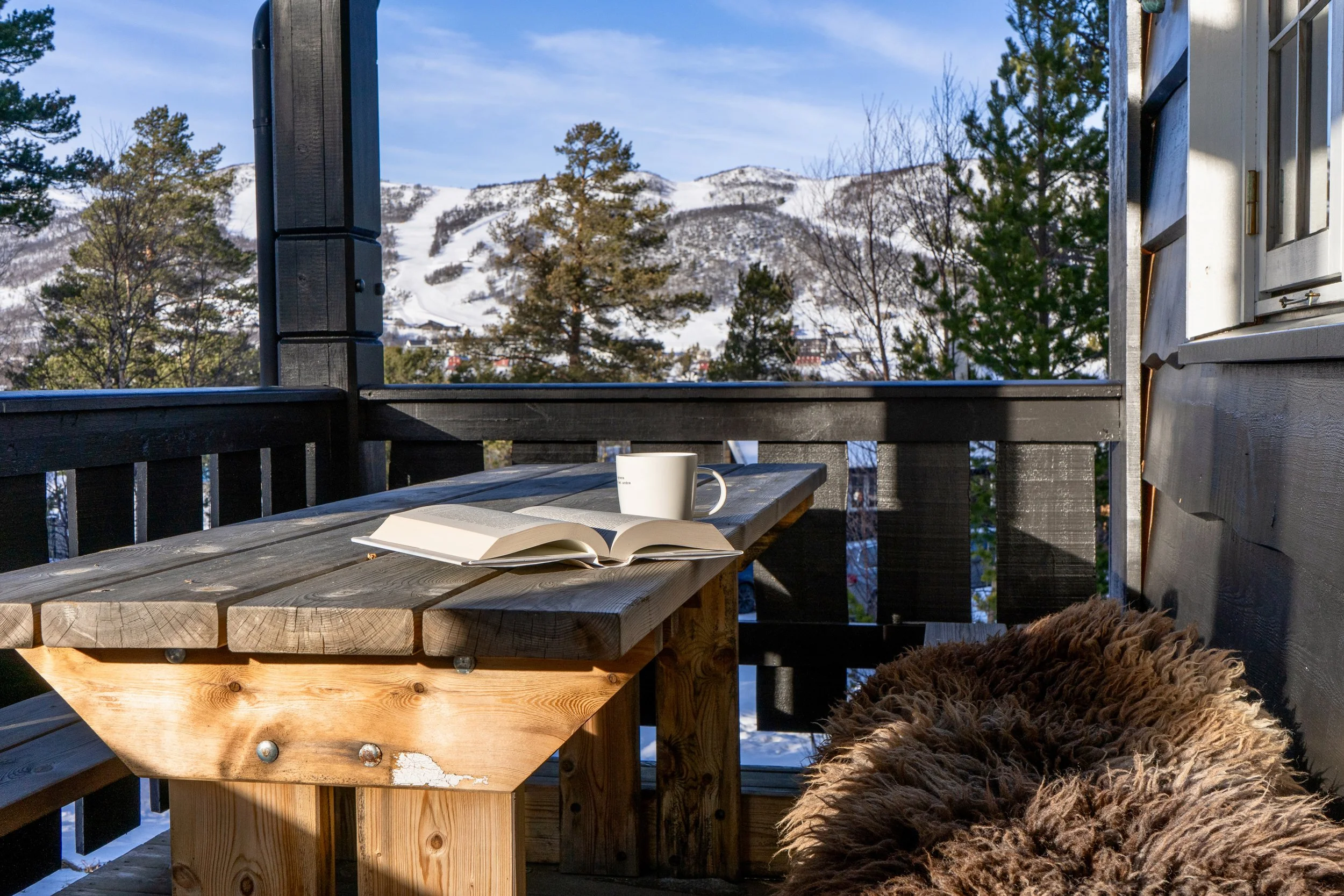 Bok, kaffekopp og saueskinn på veranda med snødekket skibakke i bakgrunnen.