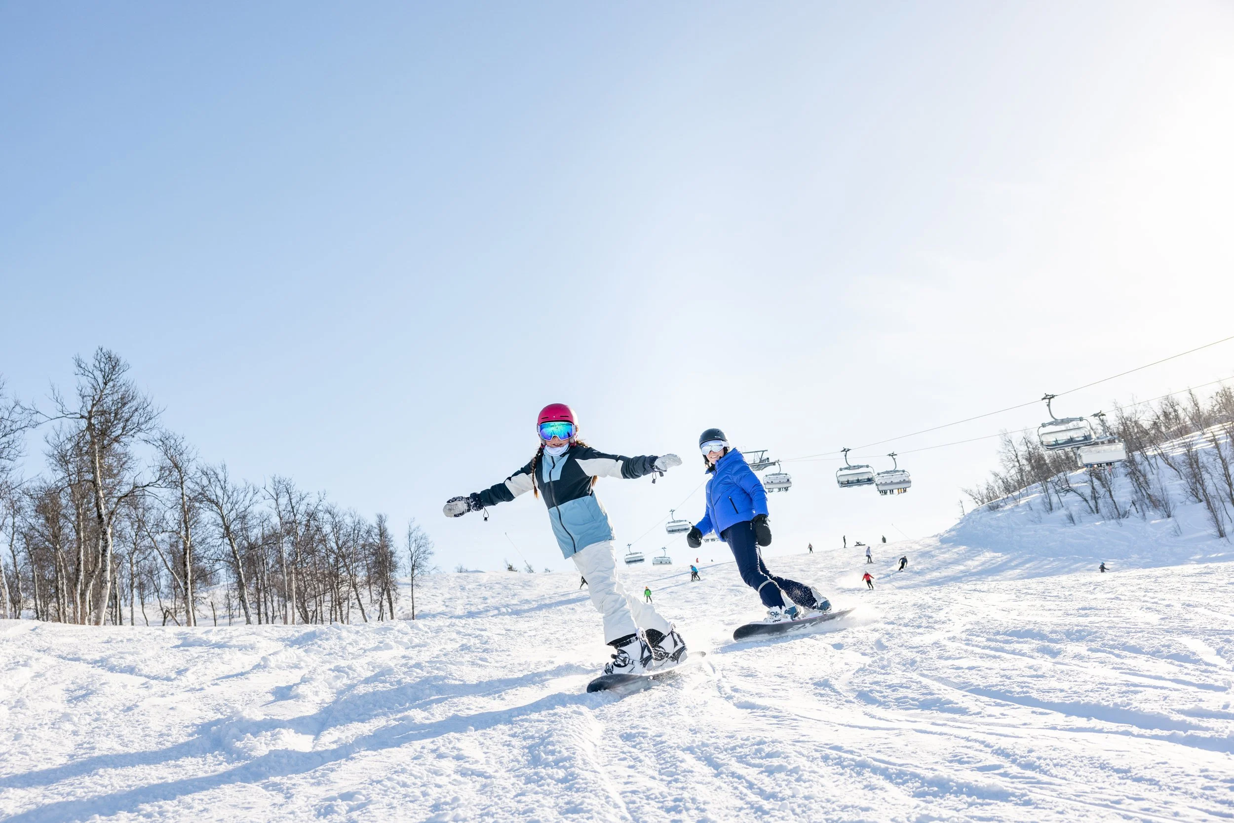 To personer står på snowboard ned skibakken med skiheis i bakgrunnen.