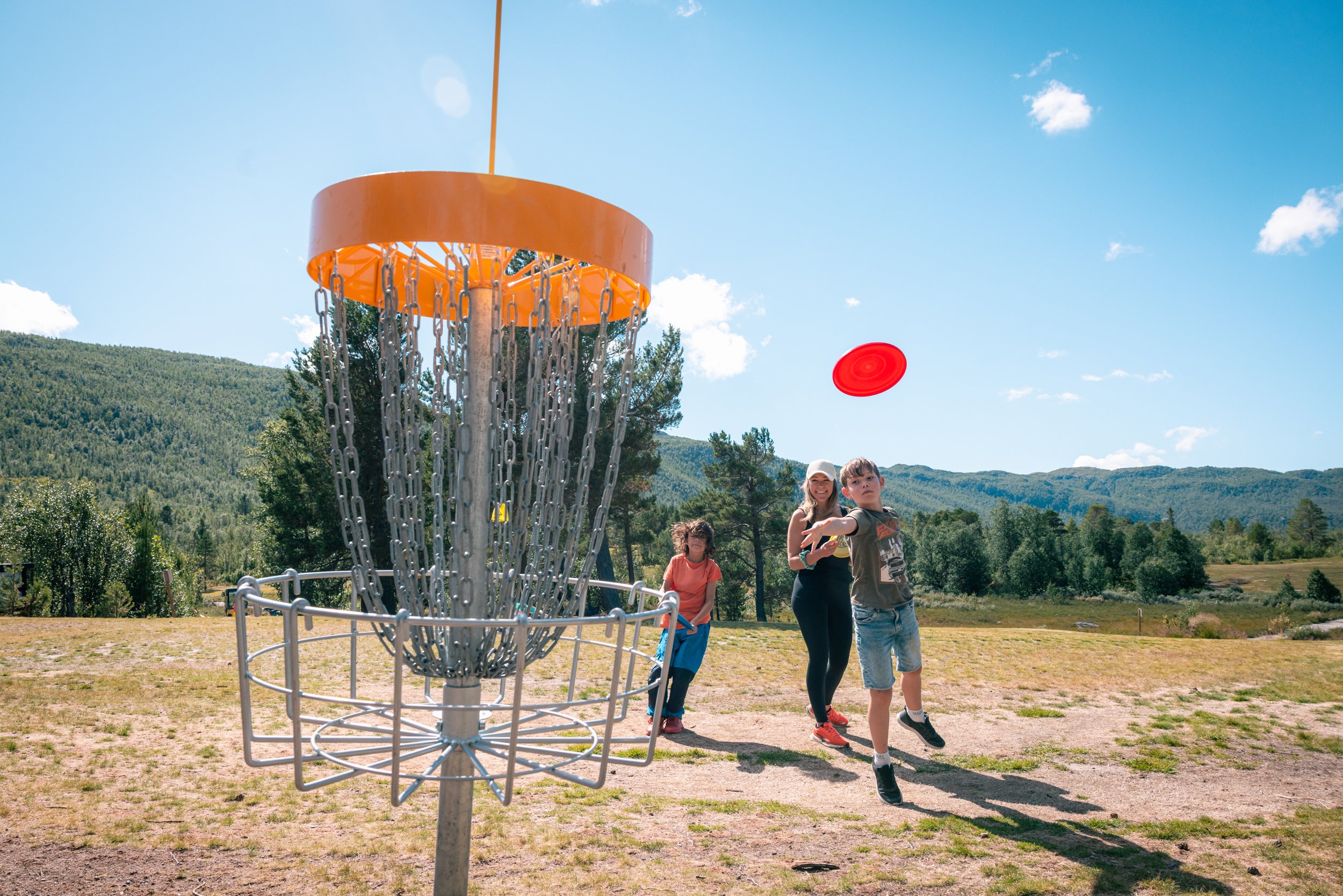Familie på tre, med en voksen og to barn, spiller frisbeegolf i sommersolen.