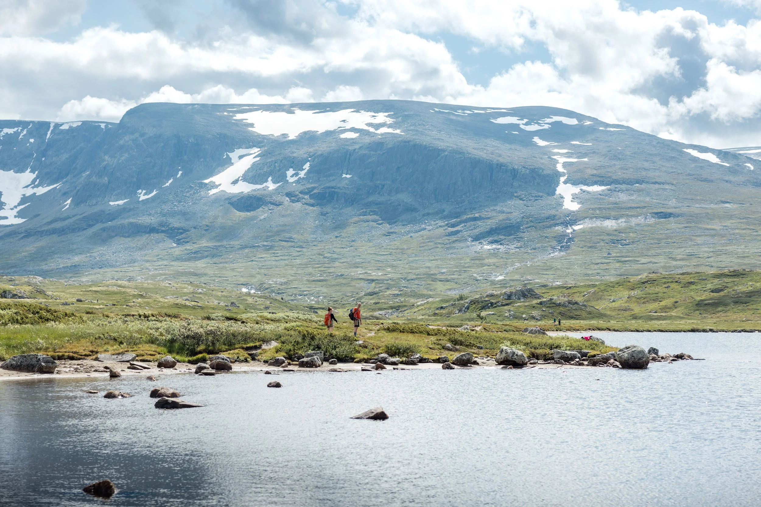 Turgåere ved fjellvann med høye fjell i bakgrunnen.